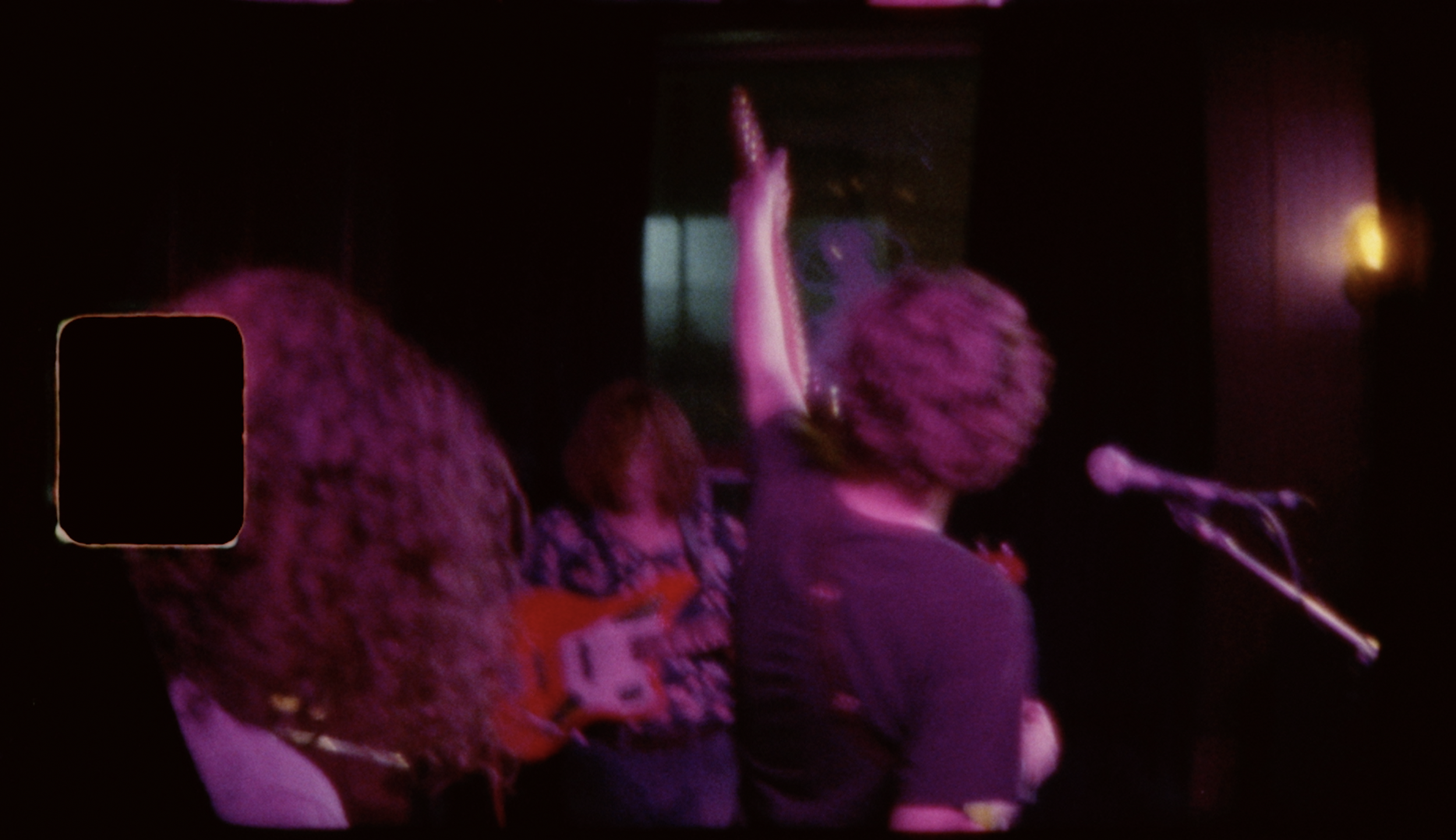 People on stage performing with a guitar and a microphone in a dark, purple-lit setting.