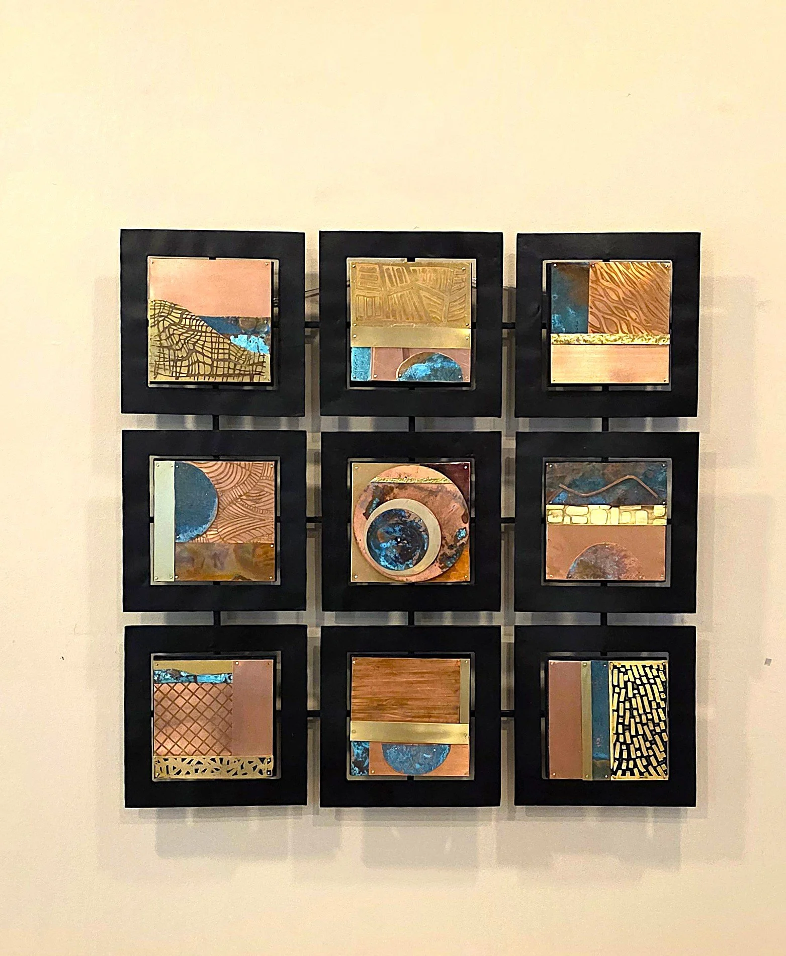 A copper wall-mounted art installation consisting of twelve rectangular metal panels, each with abstract and geometric patterns in various colors, framed in black and arranged in a 4x3 grid.
