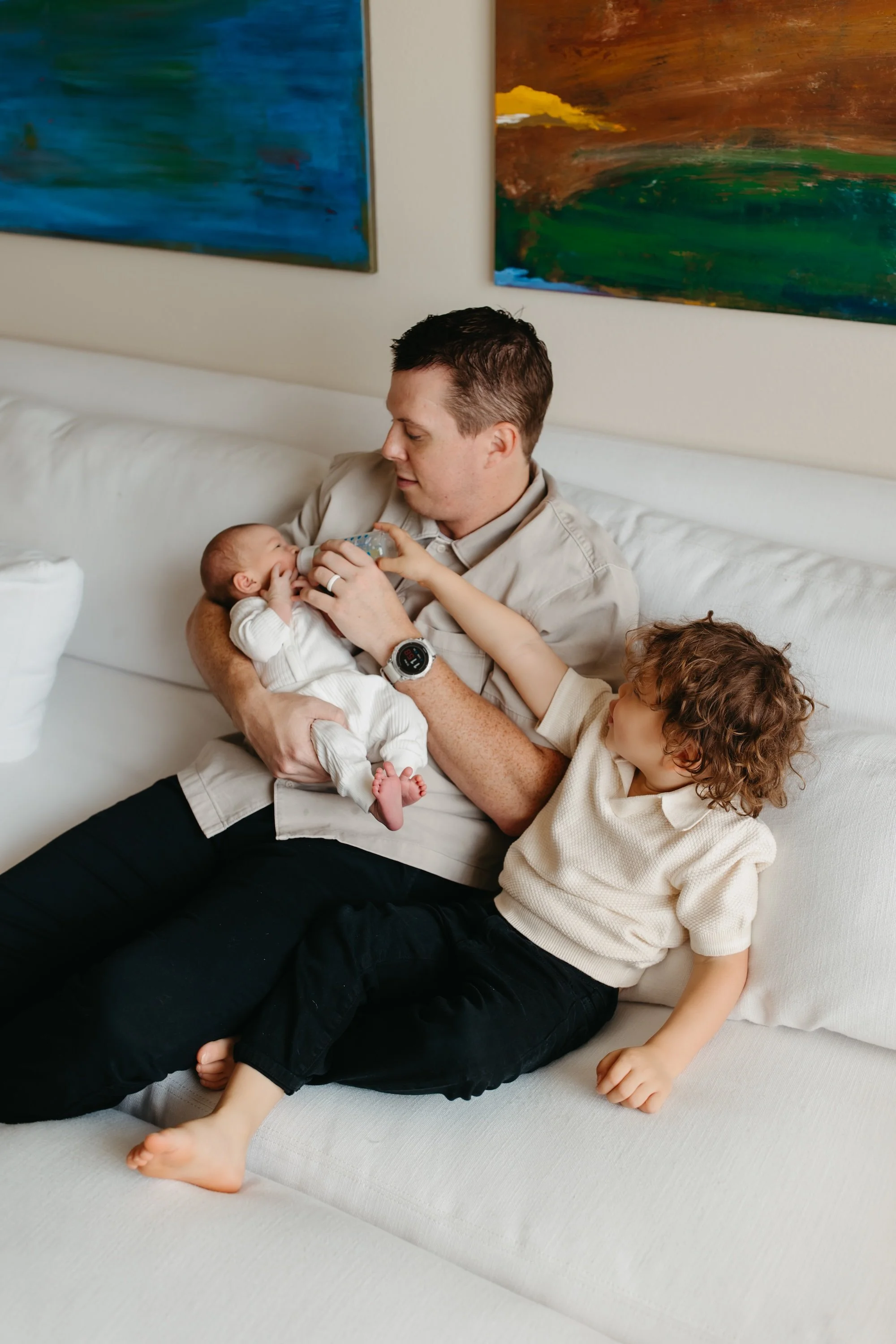 A brother helping dad feed his new baby sister during newborn photo session