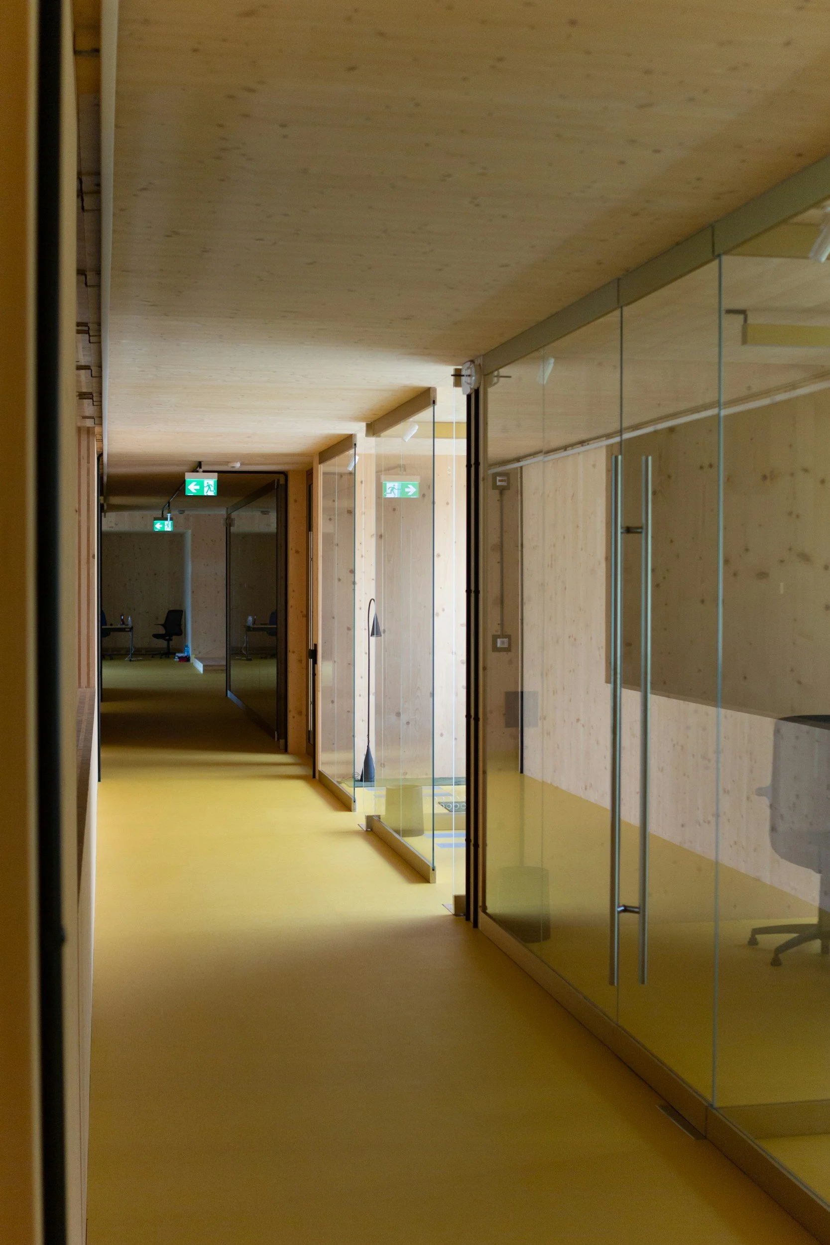 A hallway with wooden walls and ceiling, yellow flooring, glass partitions, and green exit signs.