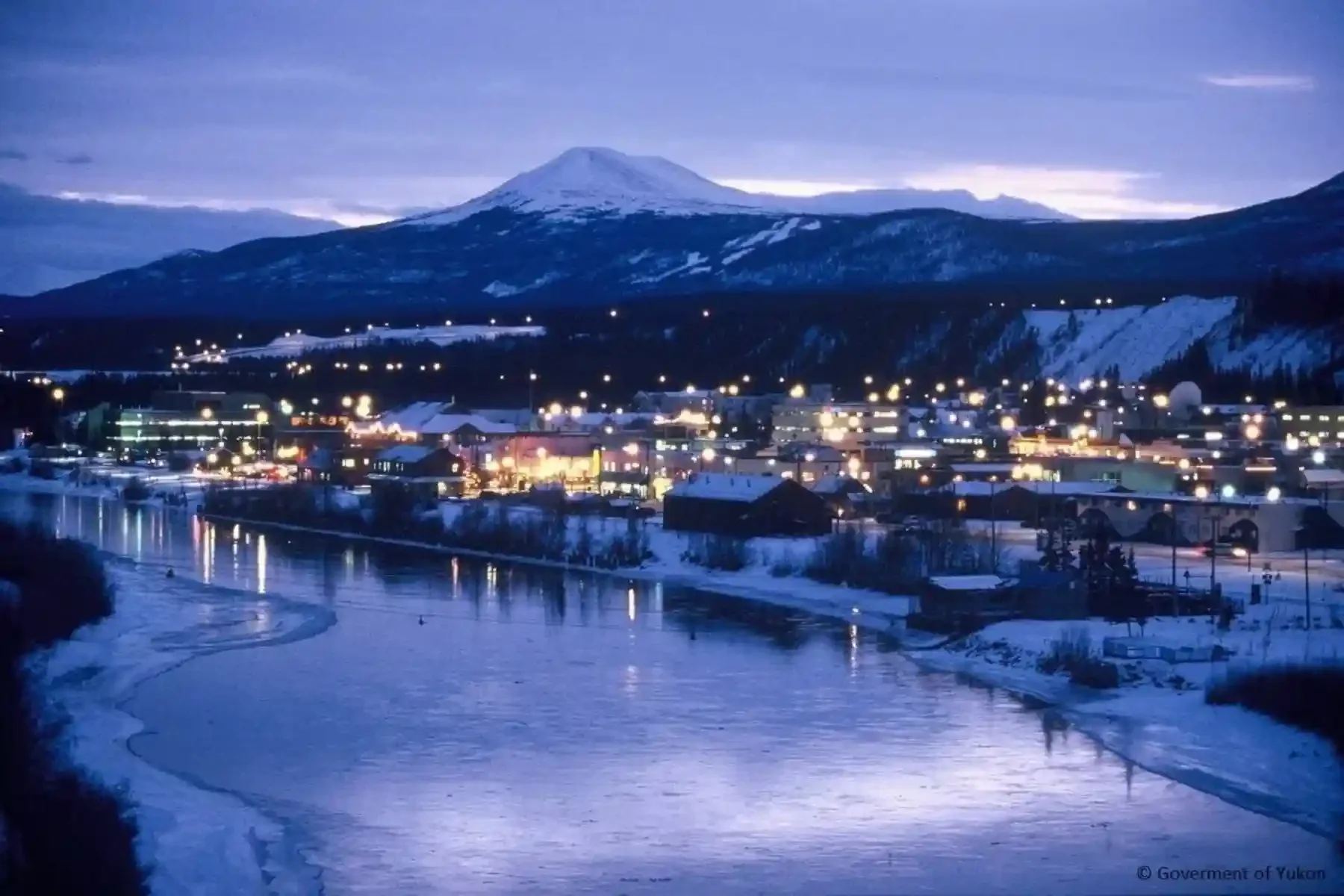 A snowy town at dusk with a river in the foreground, illuminated buildings, and a mountain in the background.