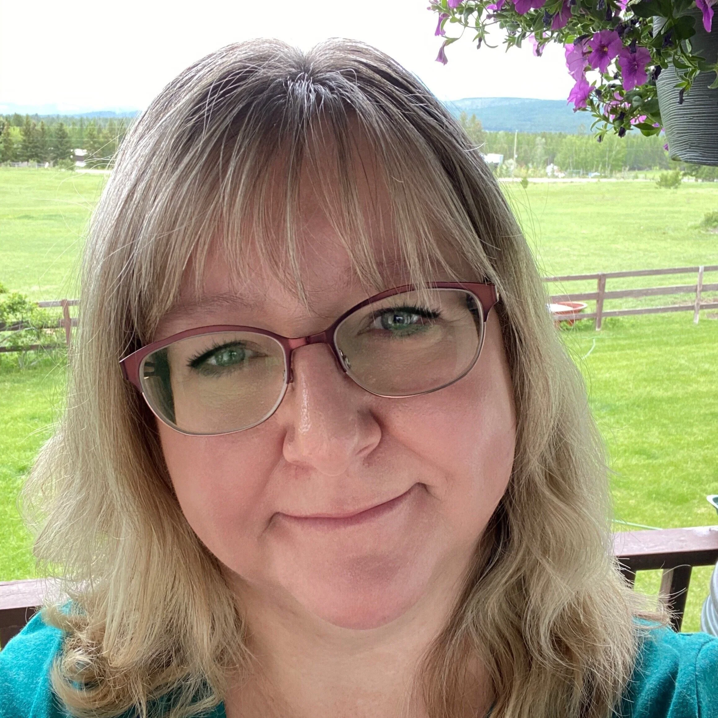 Close-up of a woman with blonde hair, glasses with pink frames, smiling, outdoors with green fields, trees, and mountains in the background.