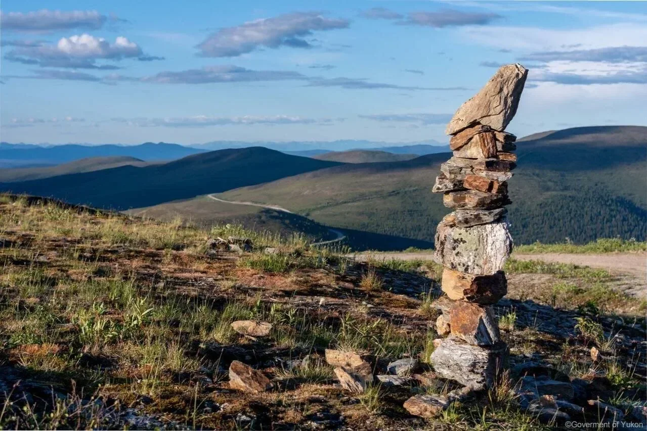 Stack of flat rocks balanced vertically in a grassy mountainous landscape with blue sky and clouds.