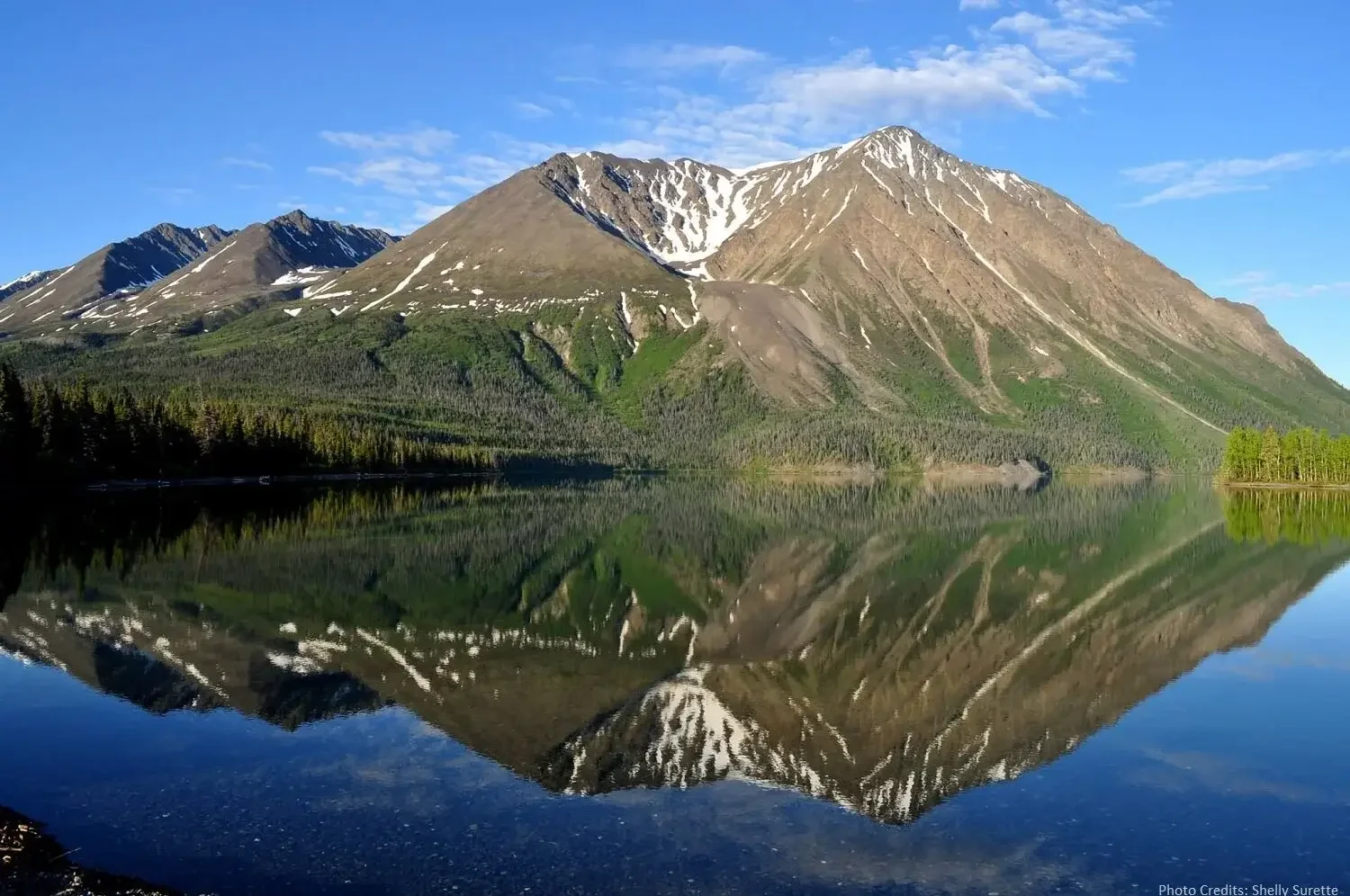 A mountain with some snow patches reflecting in a calm lake surrounded by green trees.