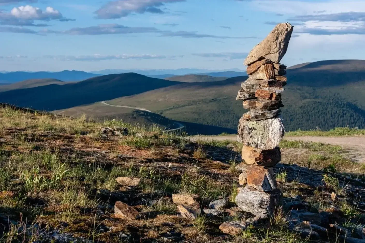 Stacked rocks on a grassy hillside with mountains and a partly cloudy sky in the background.