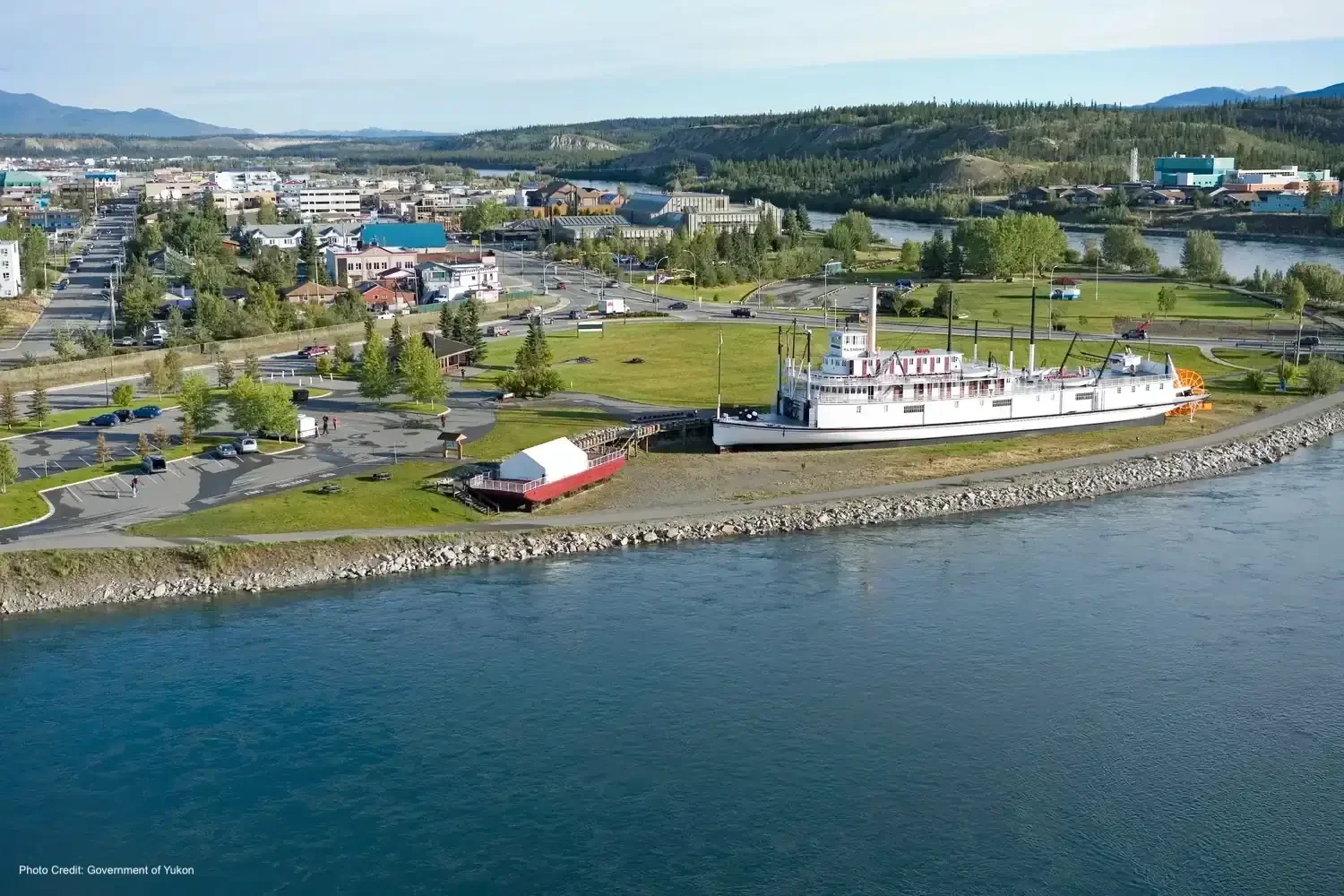 Aerial view of a waterfront park with a historic ship museum and parking lot, surrounded by trees, buildings, and mountains in the background.