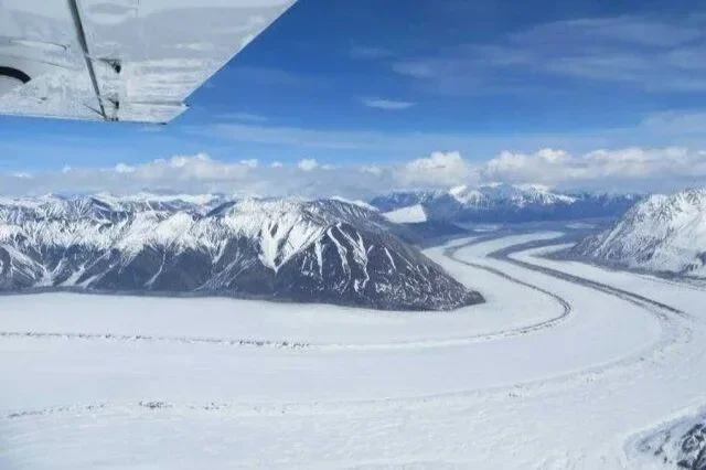 Aerial view of snow-covered mountains and a winding river or glacier in a cold, remote landscape, seen from the window of an aircraft.