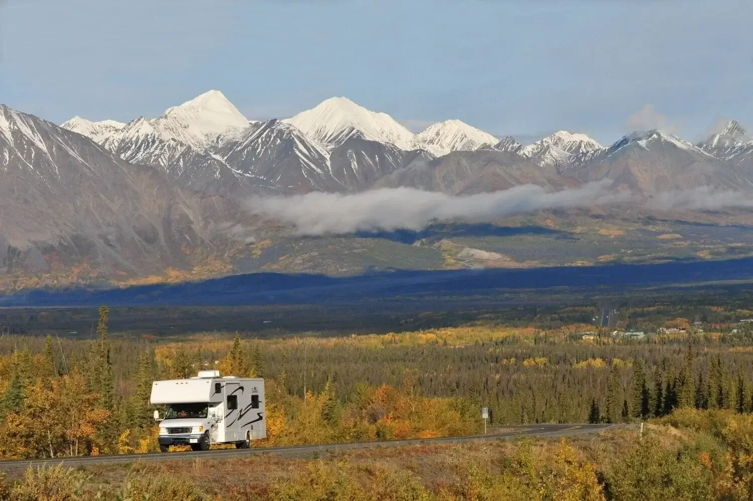 A white RV traveling on a rural road with a forest of trees displaying fall colors in the foreground, and snow-capped mountains with a layer of clouds in the background.