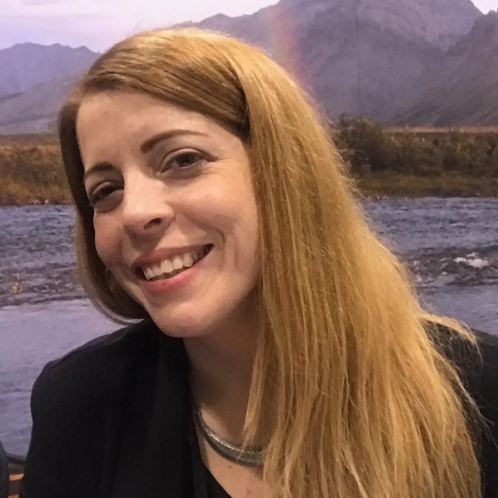 A woman with long red hair smiling in front of a scenic background of mountains, river, and purple sky.