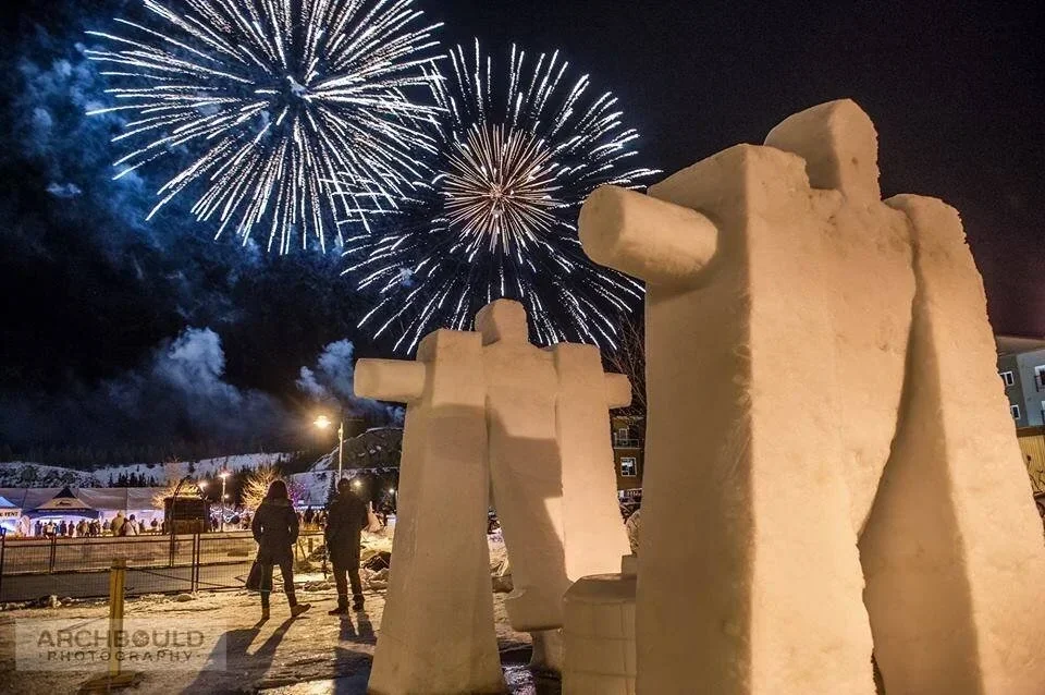 Large snow sculptures resembling human figures with fireworks lighting up the night sky in a winter outdoor setting.