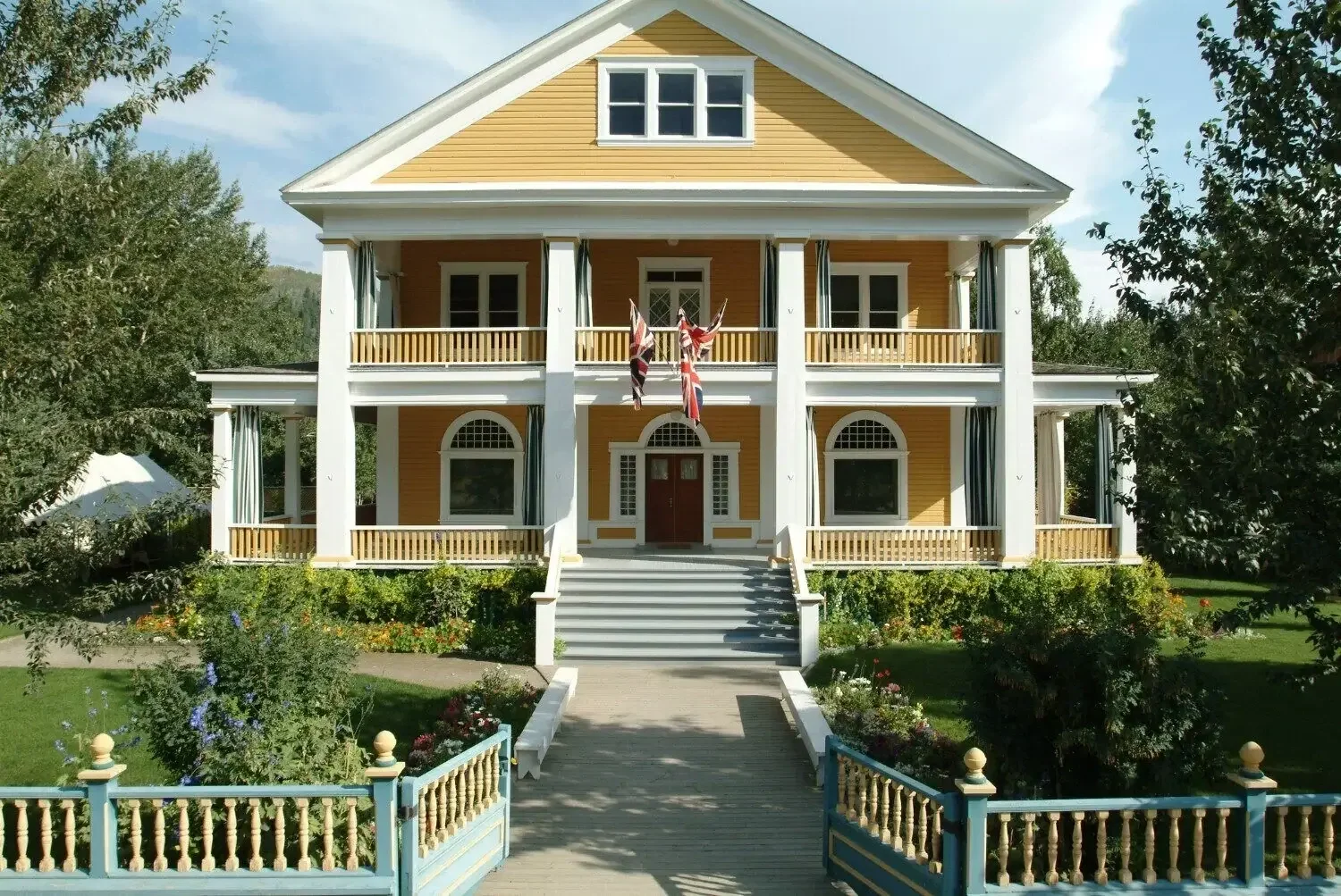 A large, yellow, three-story house with white trim and columns, red front door, multiple windows, and a front porch with Union Jack flags hanging from it. The house has a garden with flowers, trees, and a pathway leading to the steps.