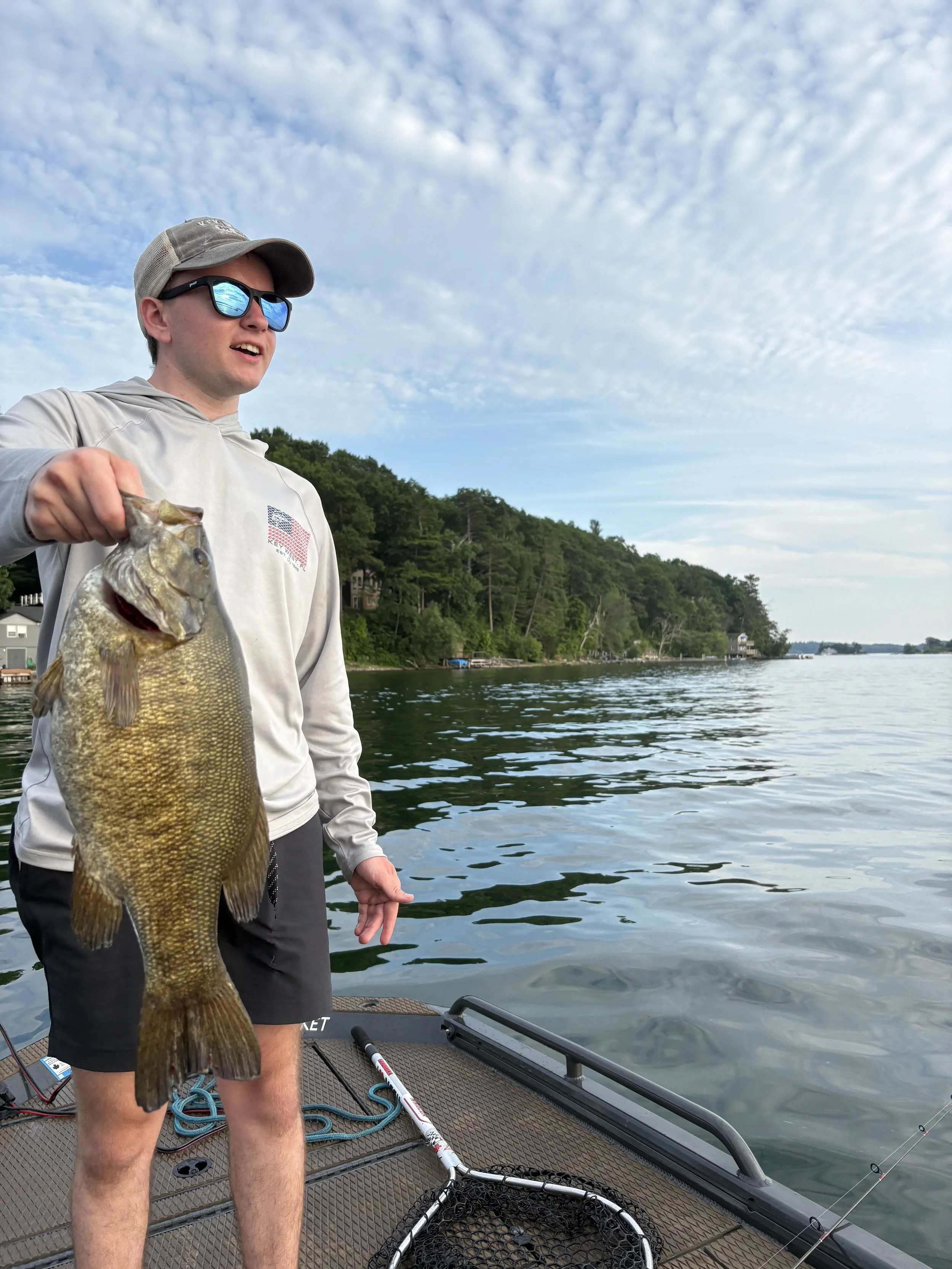 Young man on a boat holding a large fish, with a scenic lake and green hills in the background, under a partly cloudy sky.
