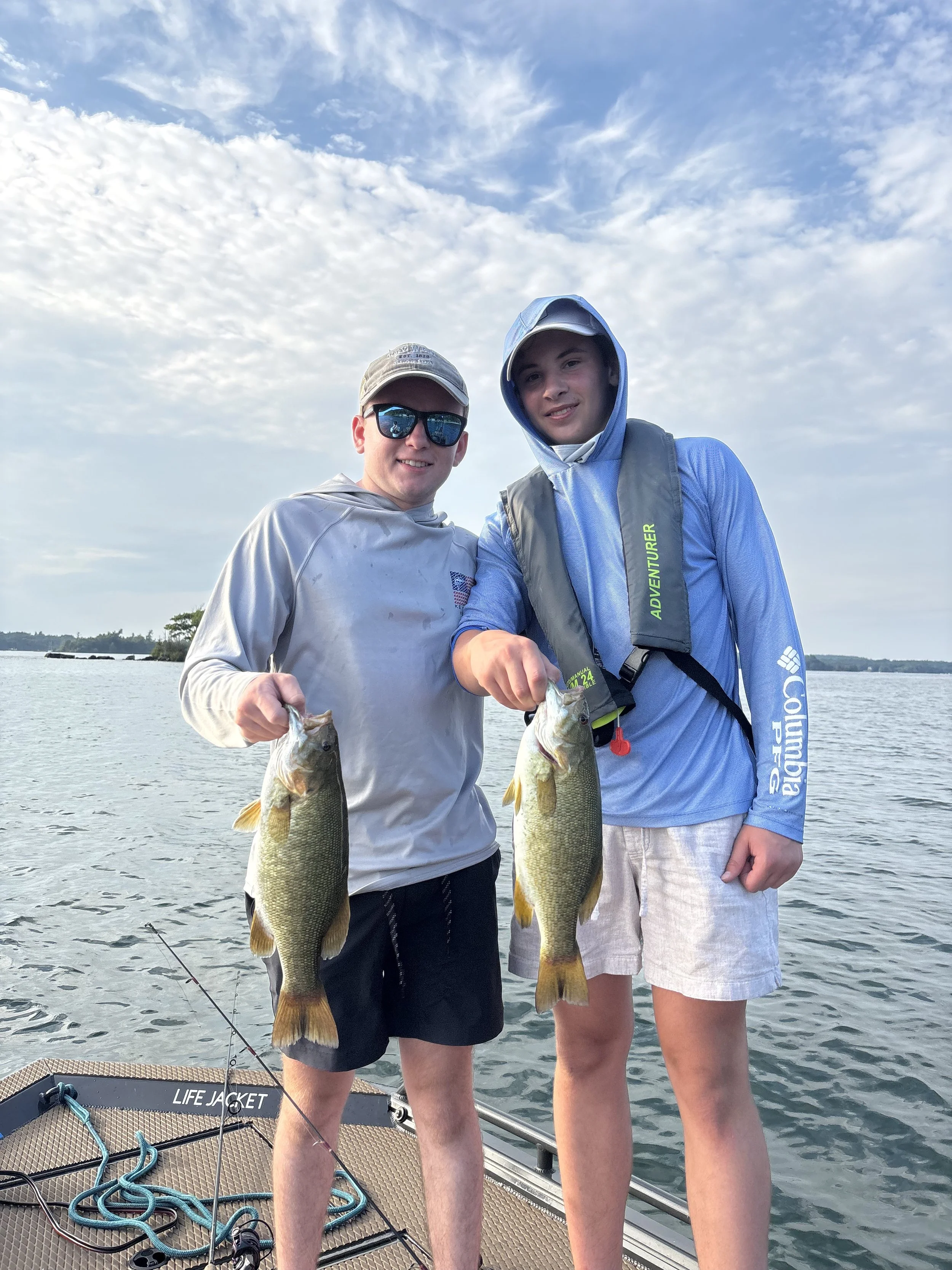 Two young men on a boat holding fish they caught during a fishing trip on a lake, with cloudy sky and water in the background.