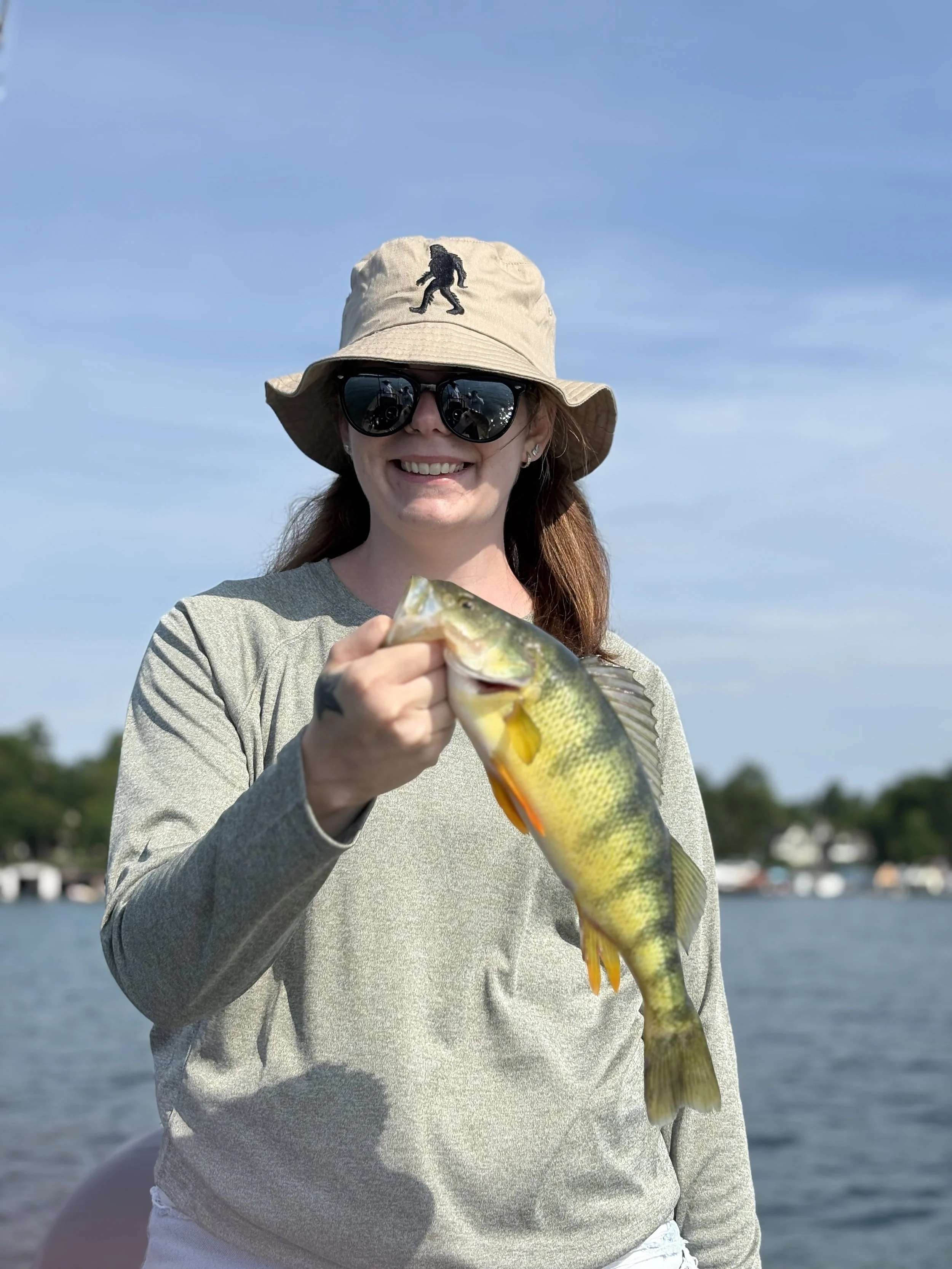 Woman holding a fish outdoors near water, wearing a beige bucket hat with a black walking figure patch, black sunglasses, and a gray long-sleeve shirt, smiling at the camera.