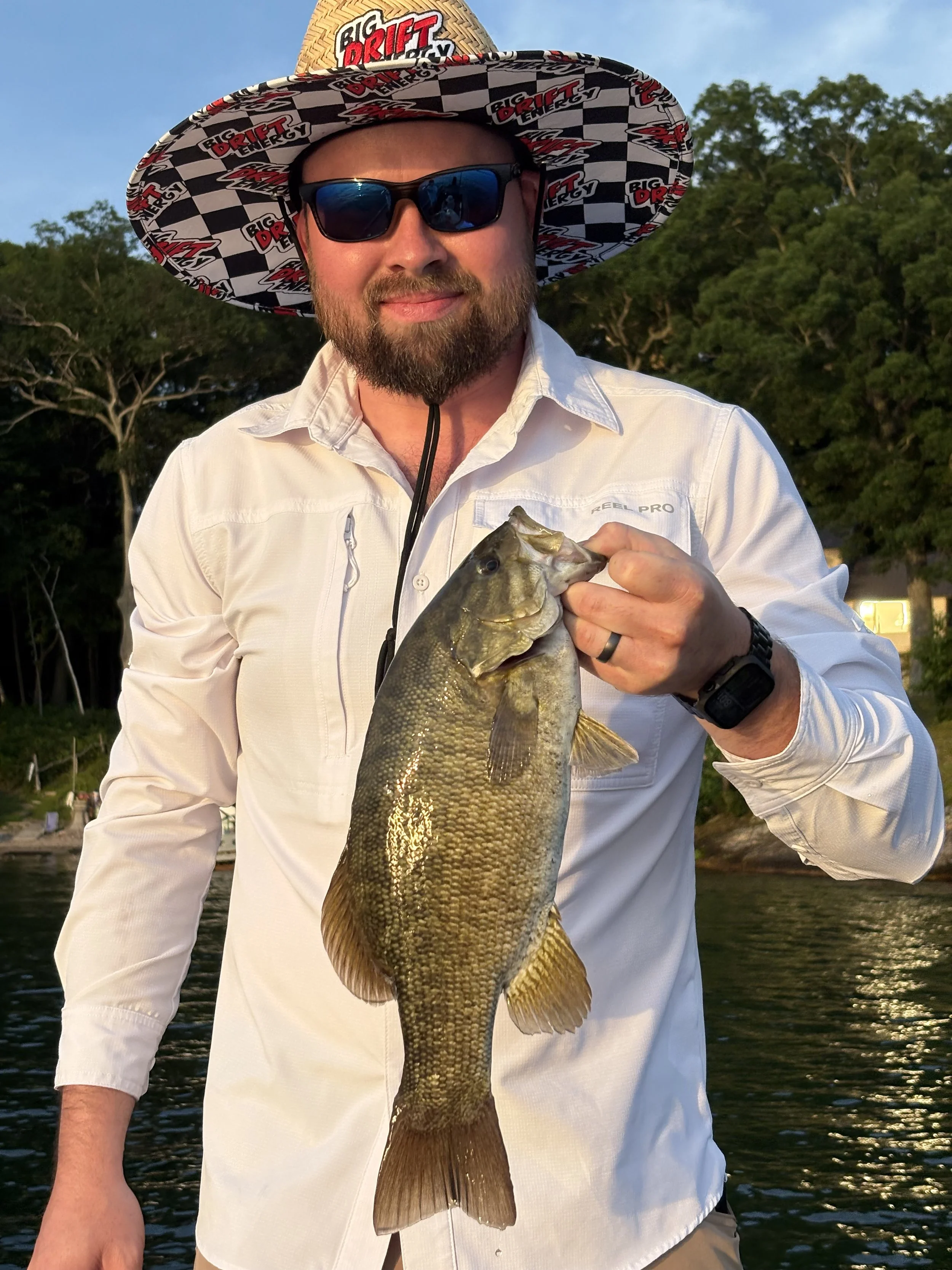 Man with beard wearing a colorful, checkered and branded Big Drift Energy hat, sunglasses, and a white fishing shirt, holding a large fish with greenish-brown scales, near a body of water surrounded by trees during sunset.