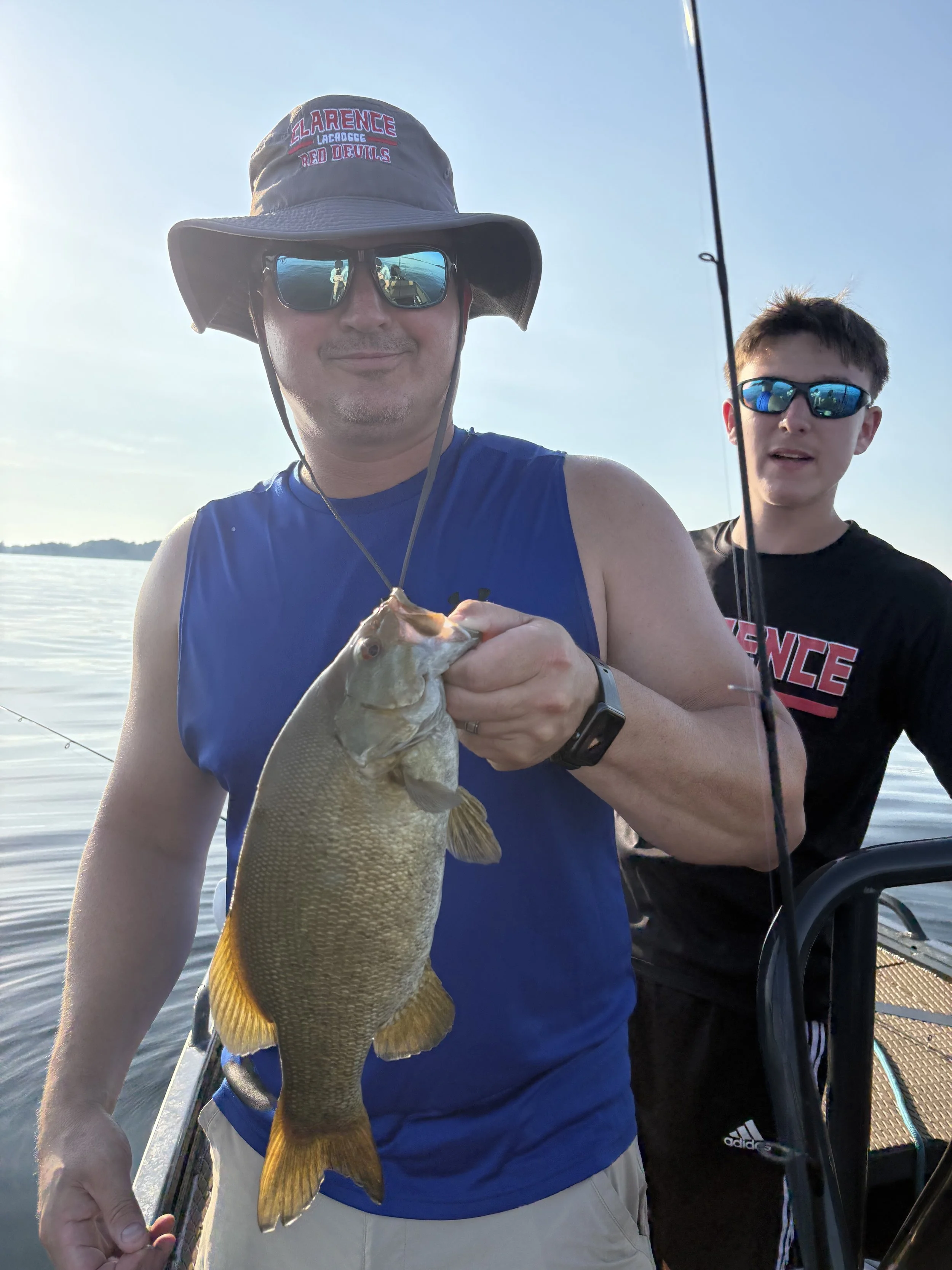 A man wearing sunglasses, a wide-brimmed hat, and a blue sleeveless shirt holding a fish he caught, with a young man in sunglasses and a black shirt standing behind him on a boat.