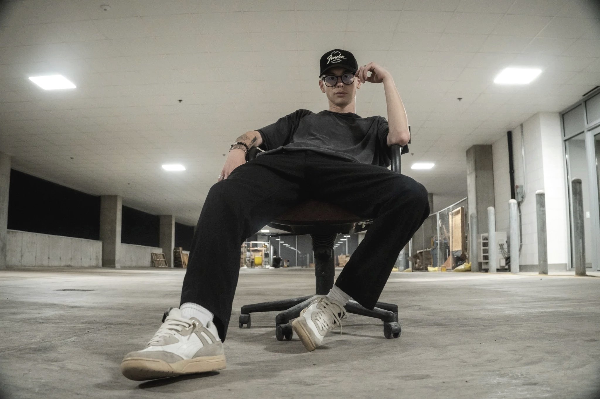 A young man with glasses and a black cap sitting in an office chair with his legs spread apart, in a large, empty indoor space with concrete floors and high ceilings.
