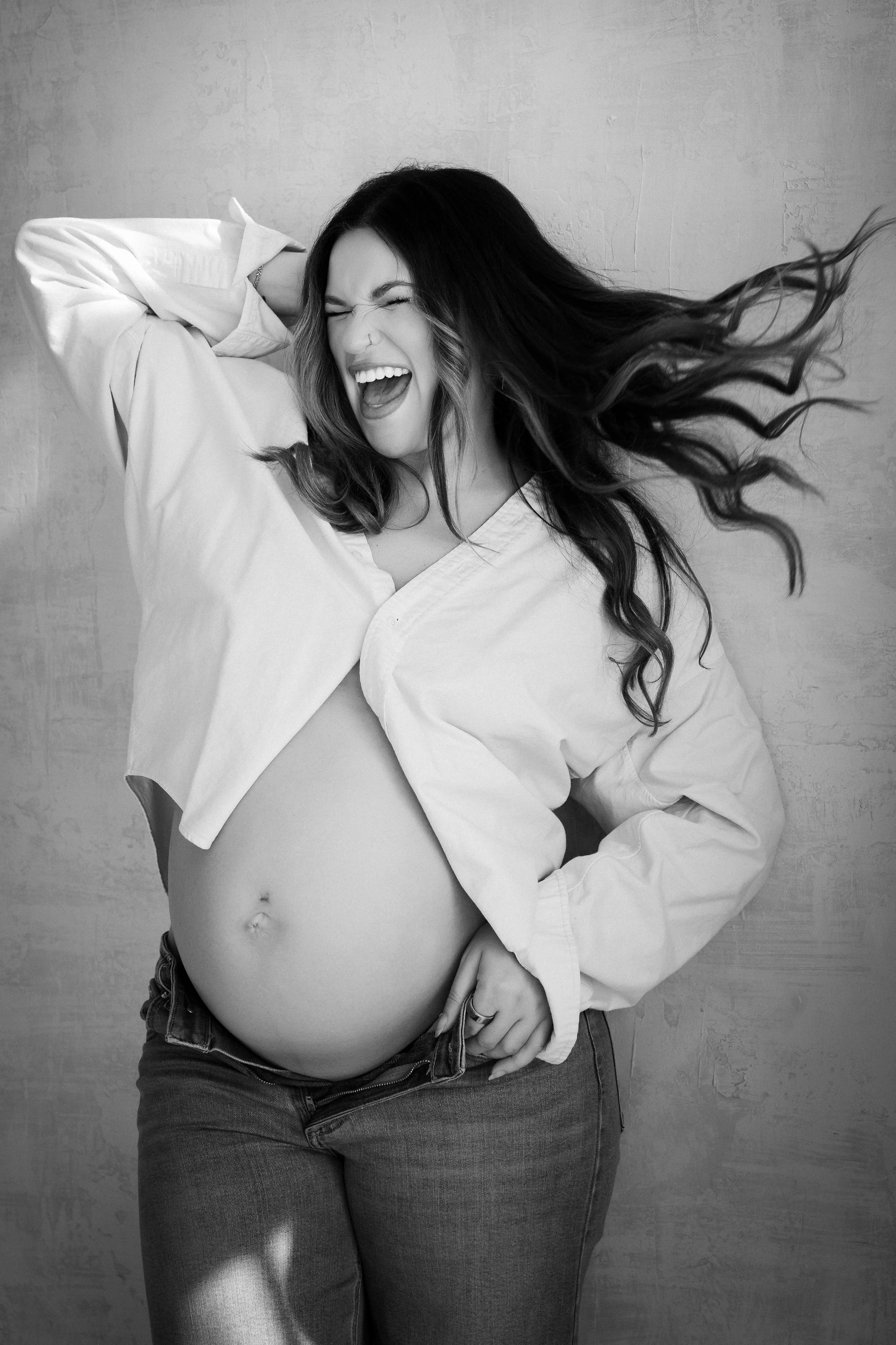 Black and white photo of a pregnant woman wearing jeans and an open white shirt, posing against a textured wall, smiling with her eyes closed.