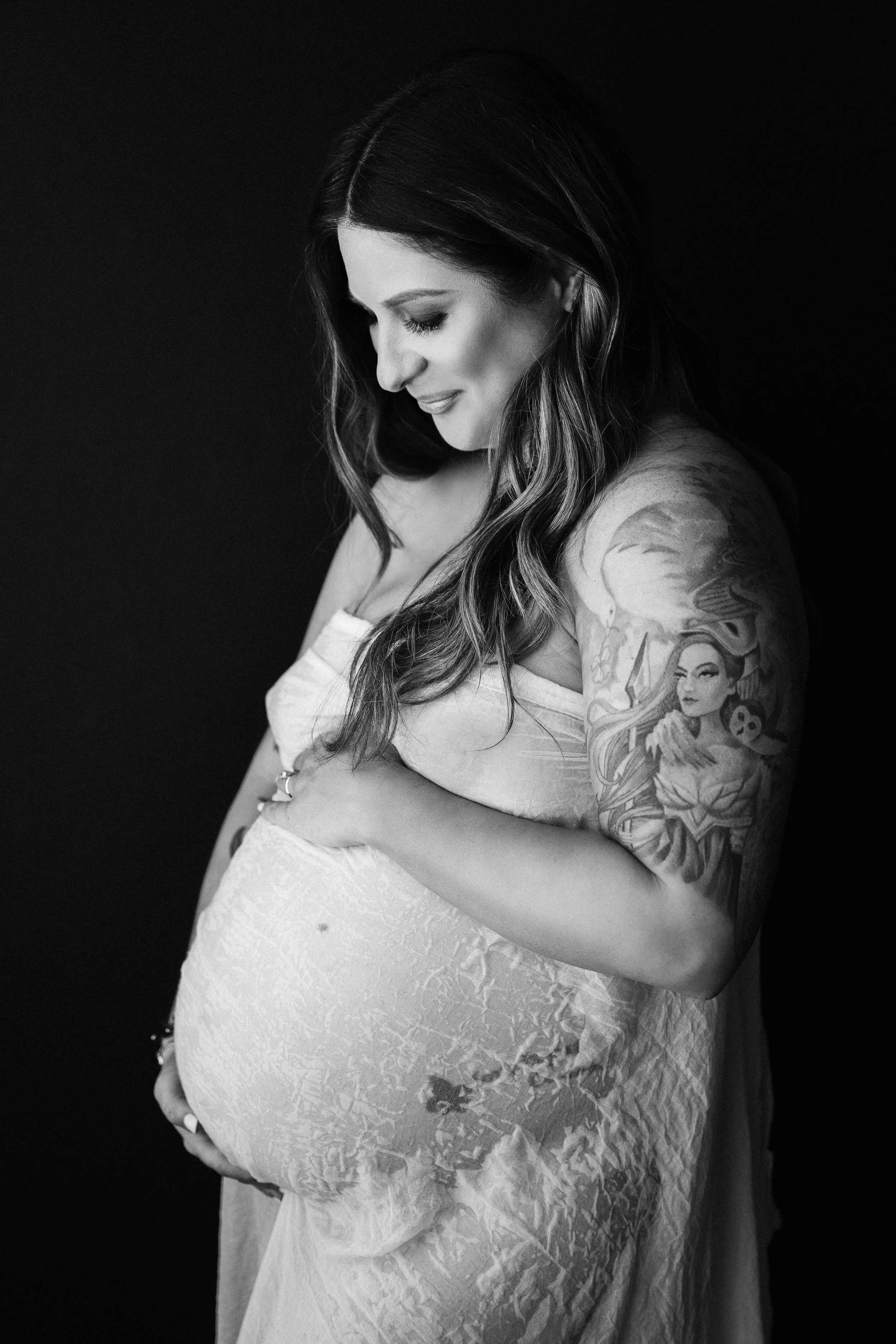 Black and white photograph of a pregnant woman with long wavy hair, looking down and smiling. She has tattoos on her upper arm featuring a woman with flowing hair, a skull, and flowers. She wears a strapless lace dress, and the background is solid black.