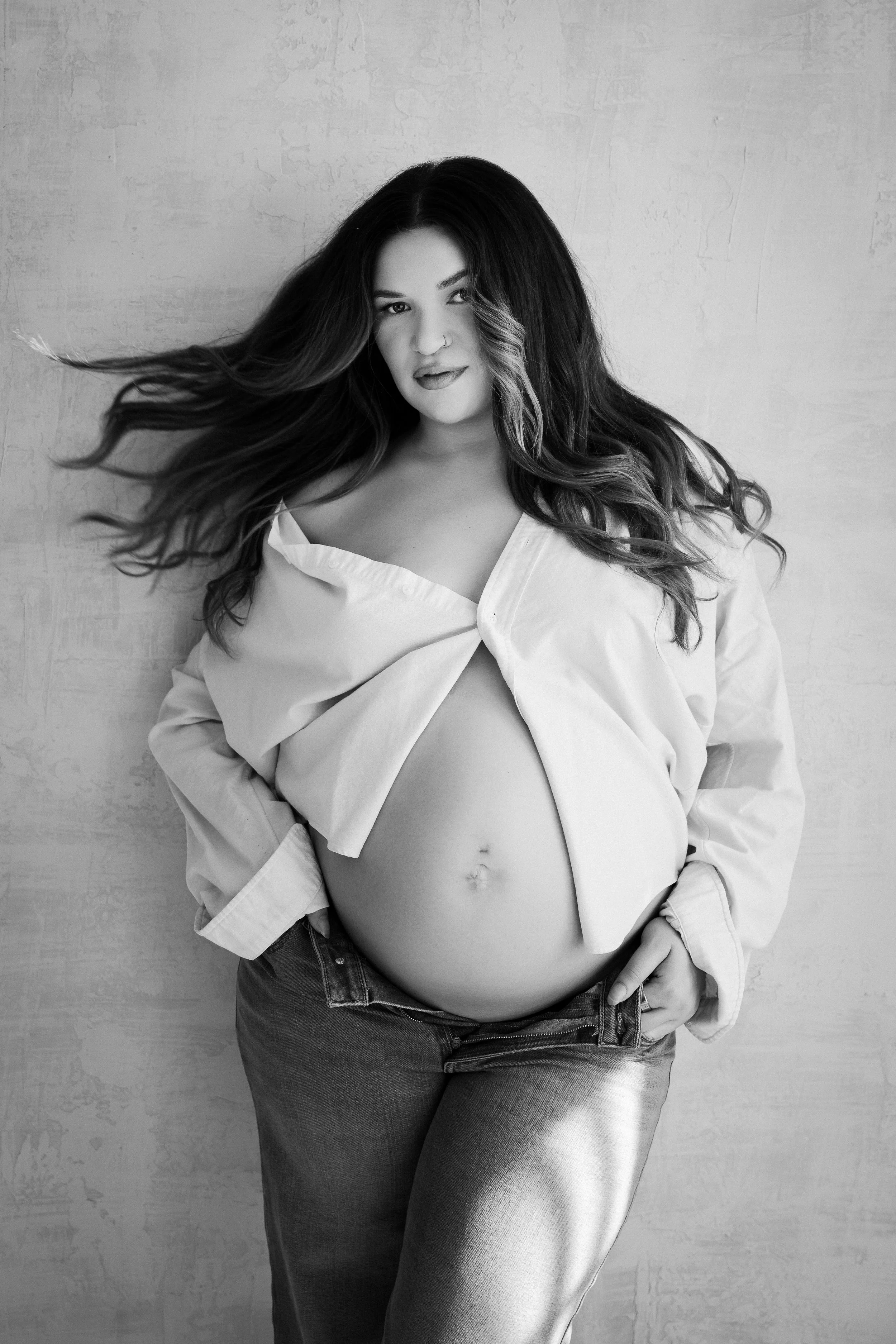 Black and white photo of a pregnant woman with long, wavy hair, wearing a partially open shirt and jeans, standing against a textured wall.