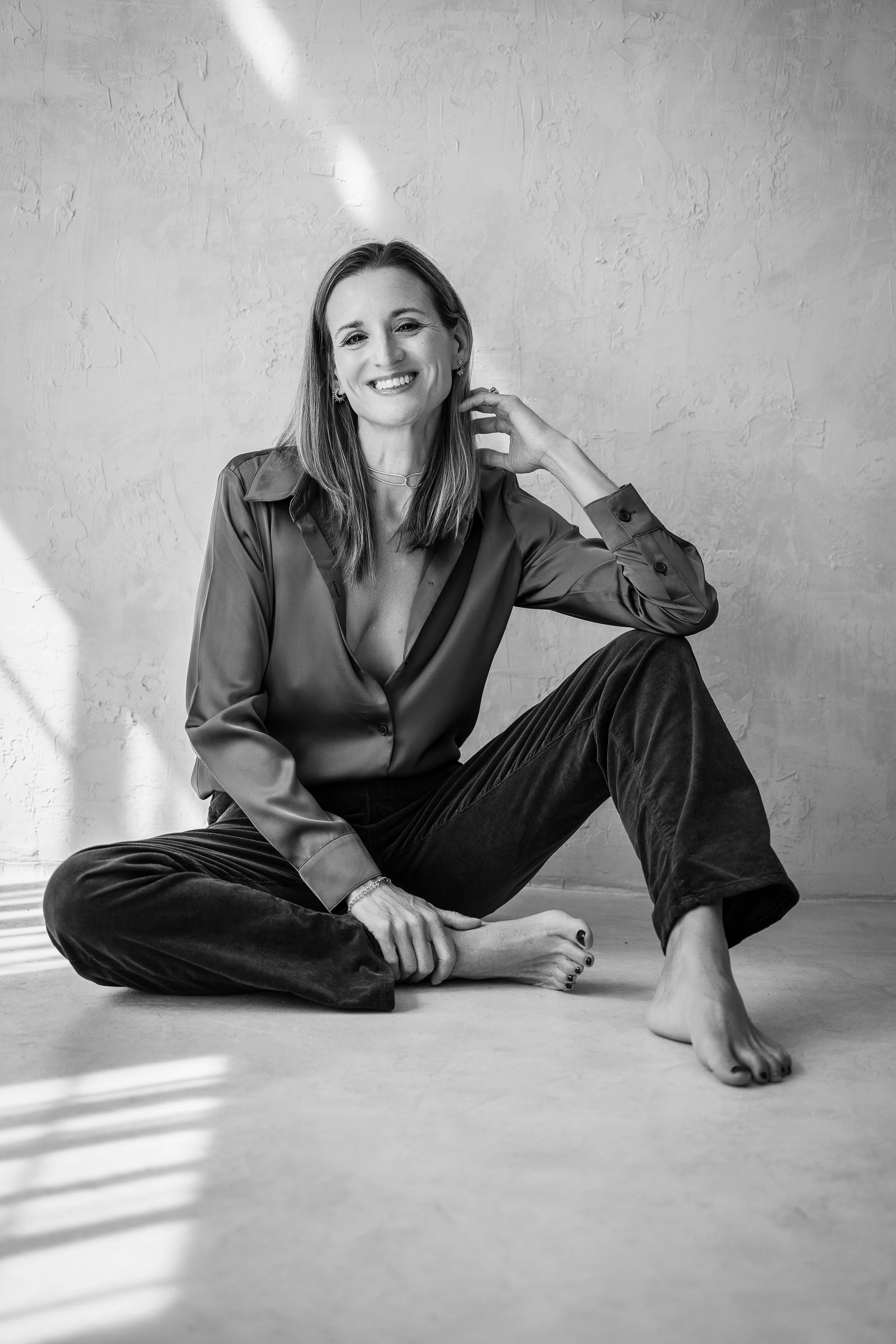Black and white photo of a woman in a blazer, sitting on a chair, with one arm on the back of the chair and one leg extended, posing confidently against a plain wall background at Chrissy Rene Studios in Round Rock, Texas