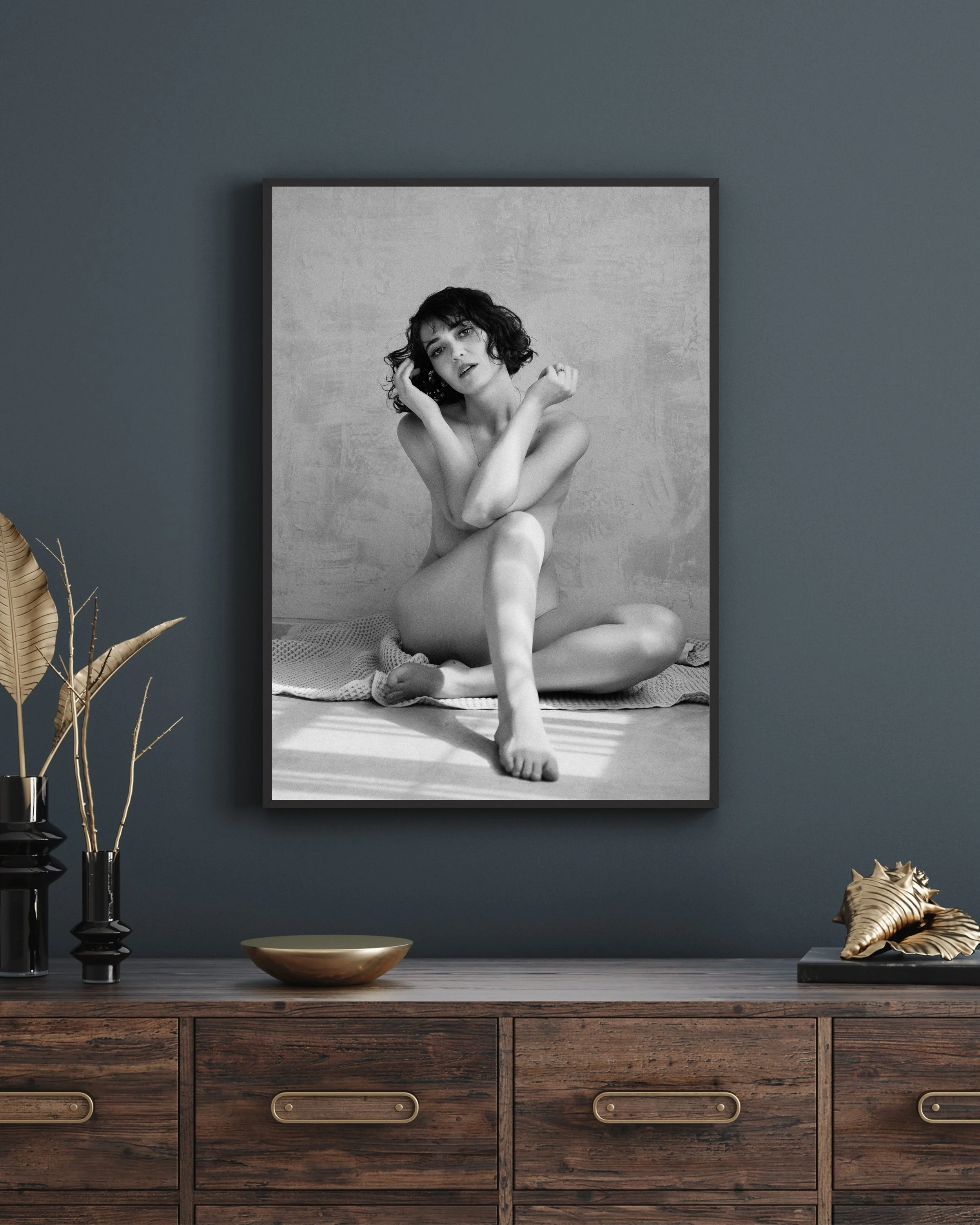A black-and-white photograph of a woman with curly hair sitting with her legs crossed, touching her face with her right hand, and looking into the camera from her boudoir session at Chrissy René Studios in Round Rock, Texas