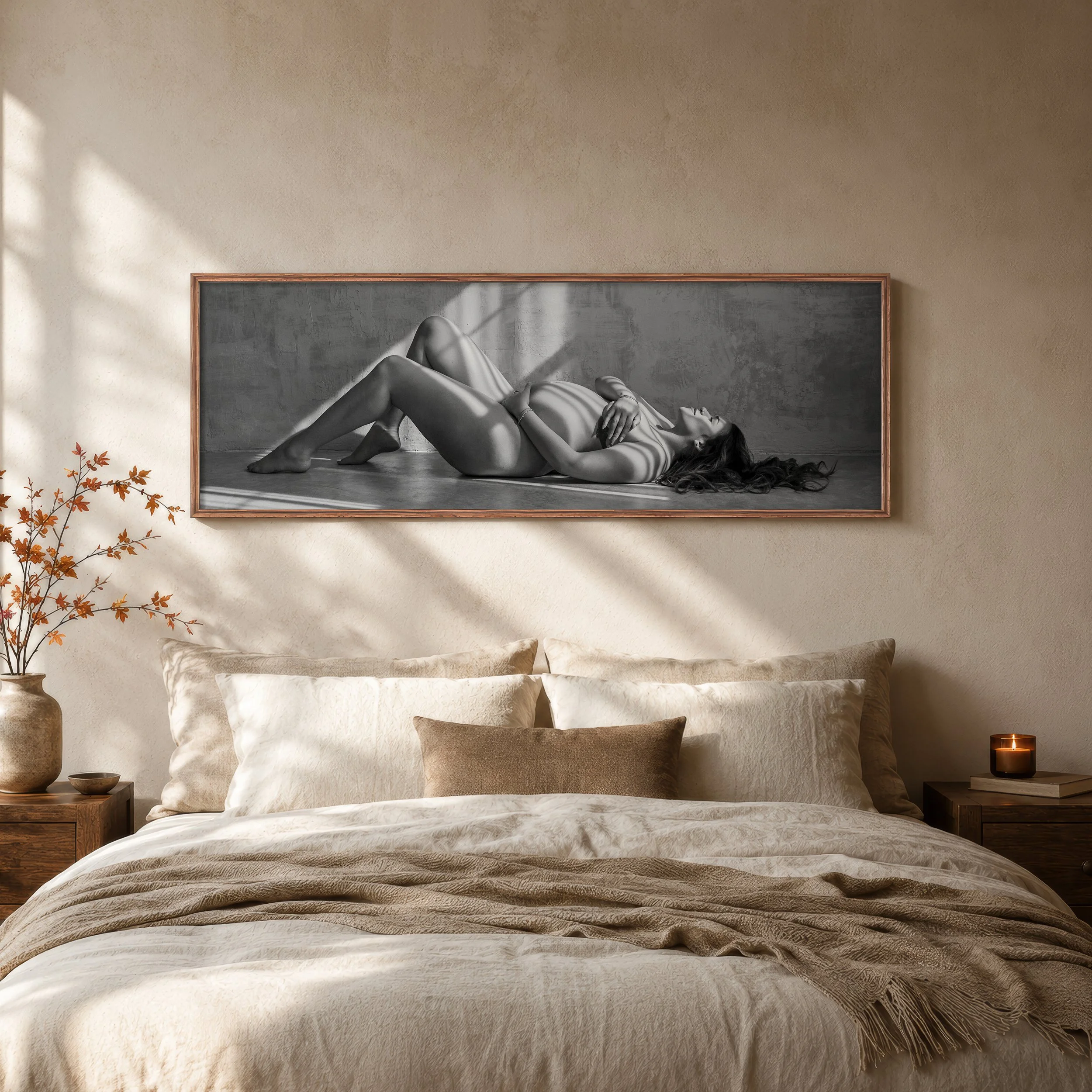 A bedroom with a large framed black and white photograph of a nude woman lying on the floor, leaning against a textured wall. The bed is made with neutral-colored bedding, and there are decorative pillows. A wooden nightstand with a small plant and a candle is visible on the left, while a small dark wooden table with a lit candle stands on the right. A beige wall with soft shadows and a few plants with orange leaves in the corner add warmth to the room.