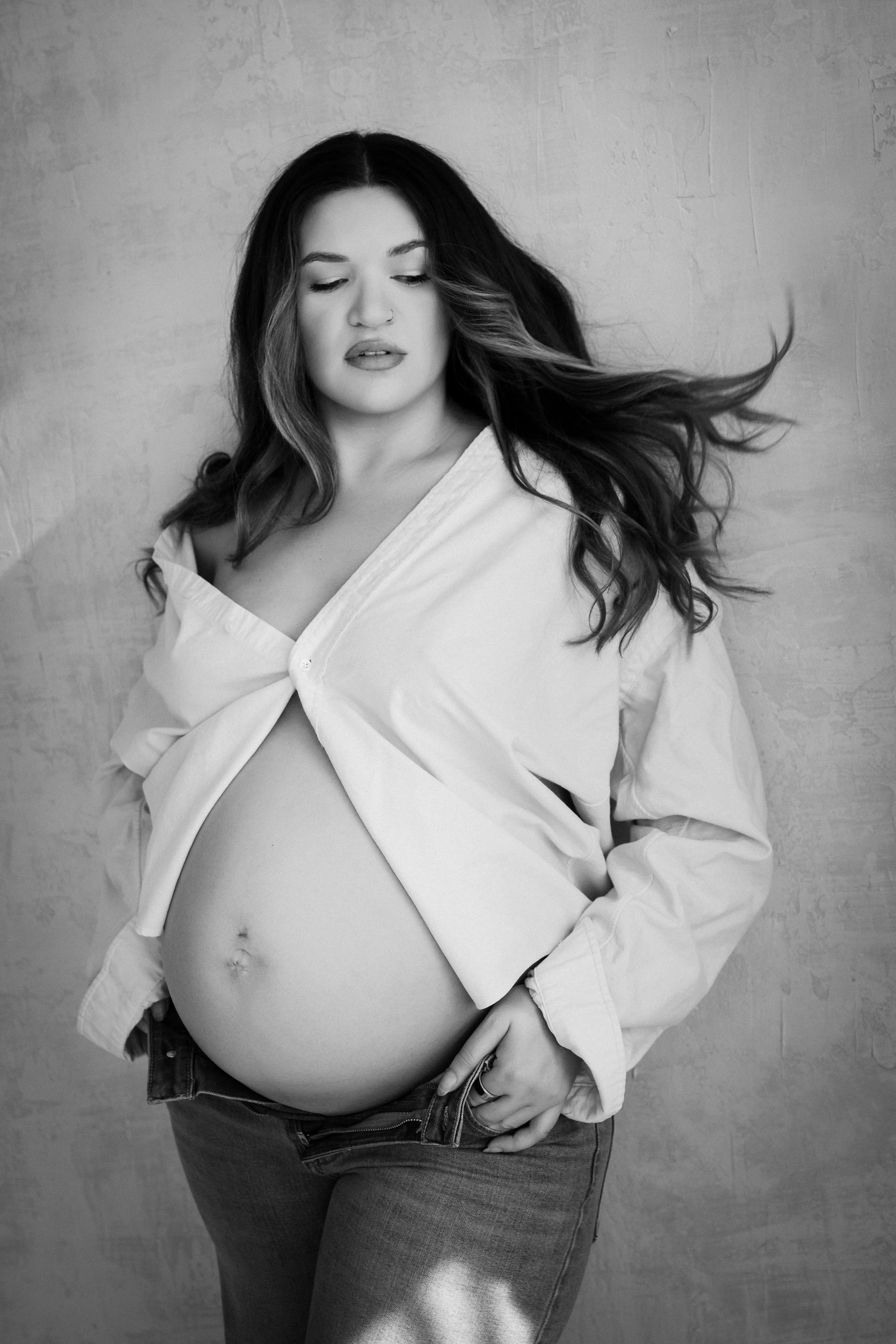 Black and white photo of a pregnant woman wearing a white shirt and jeans, against a textured wall, with her hand on her belly and her eyes closed.