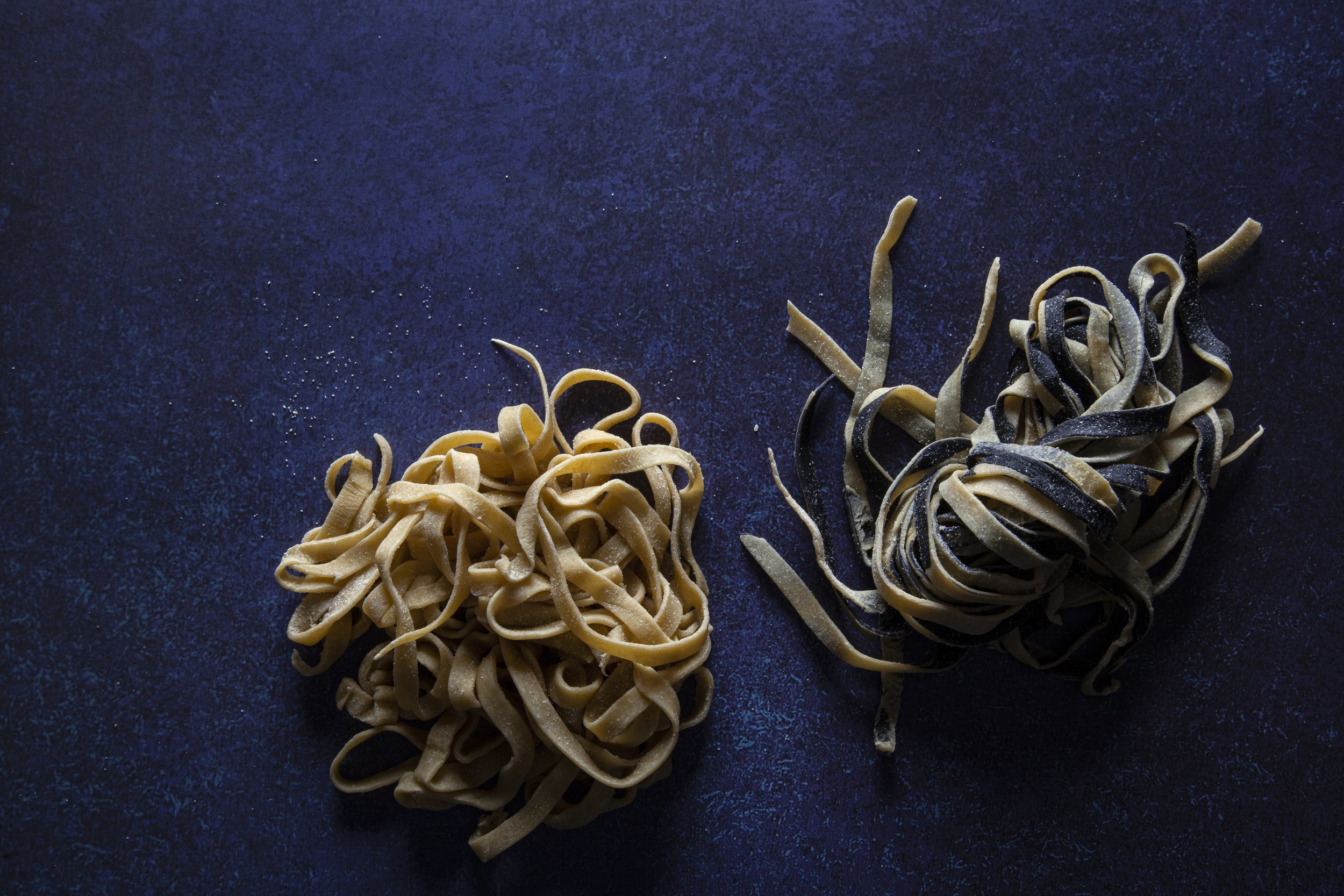 Two piles of uncooked pasta, one plain tagliatelle and the other black squid ink tagliatelle, on a dark surface.