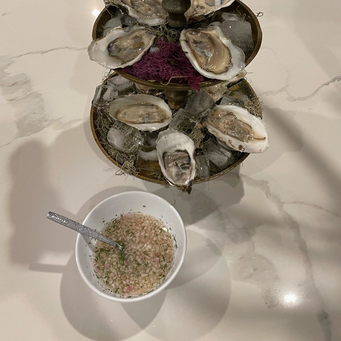 A tiered platter with raw oysters on ice, accompanied by a bowl of minced shallots and herbs in a vinegar mixture.