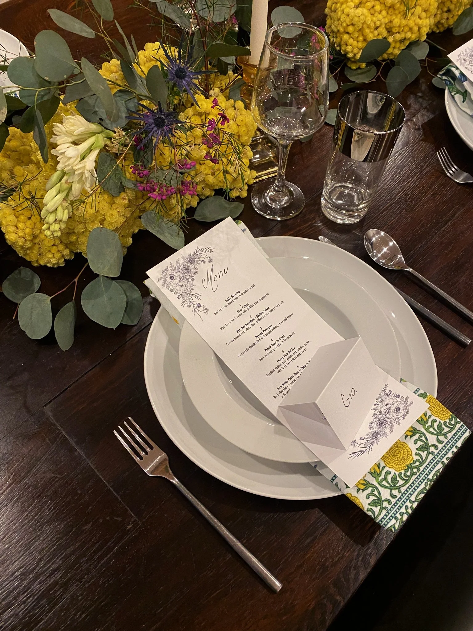 Dining table setup with a menu card on a plate, floral arrangement with yellow and white flowers, and eucalyptus leaves, accompanied by wine and water glasses, and cutlery on a dark wooden table.