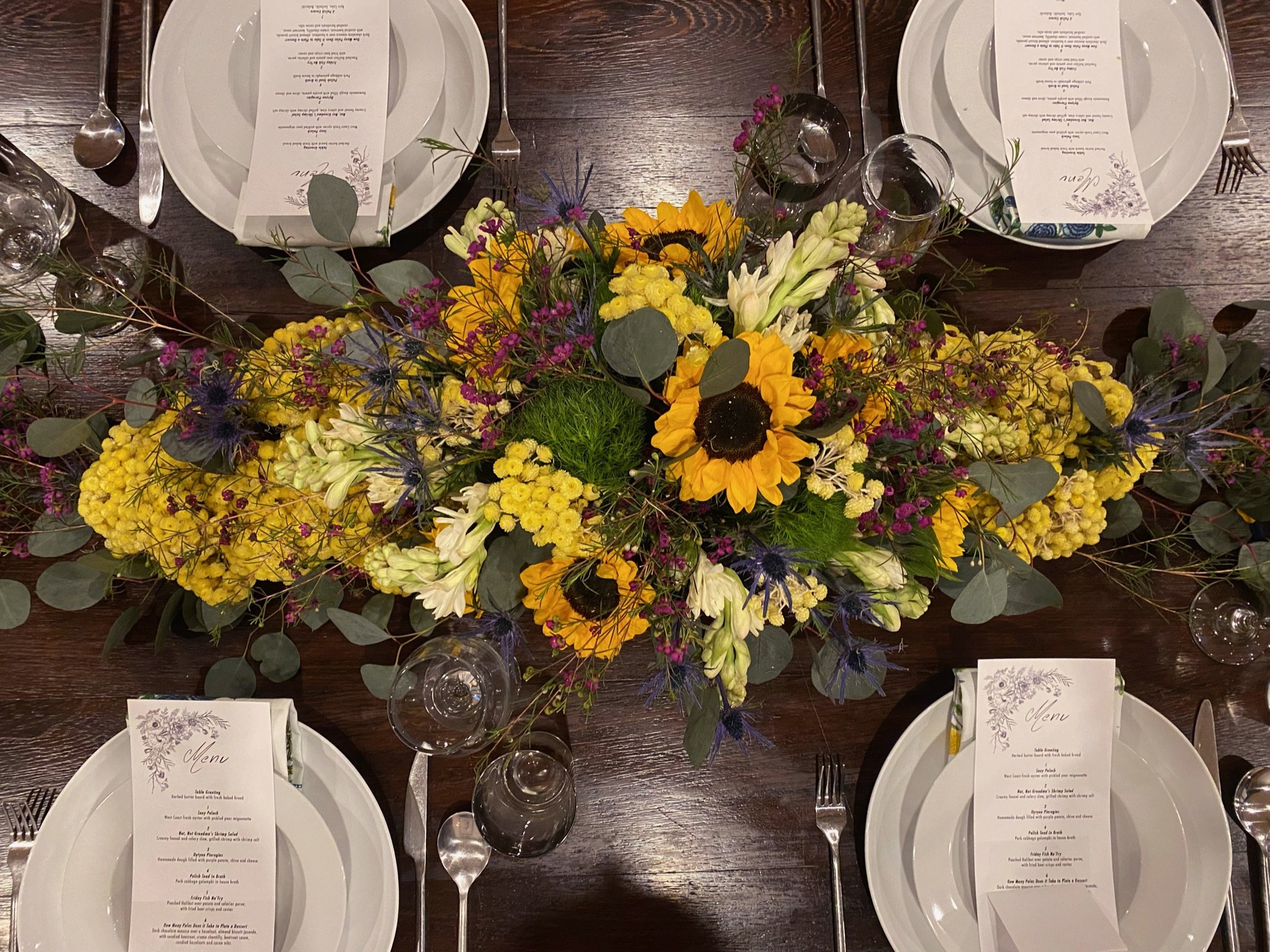 A dining table setup with elegant floral centerpiece featuring yellow sunflowers and various mixed flowers. The table includes white plates, silver cutlery, and wine glasses. Menus are placed on the plates.