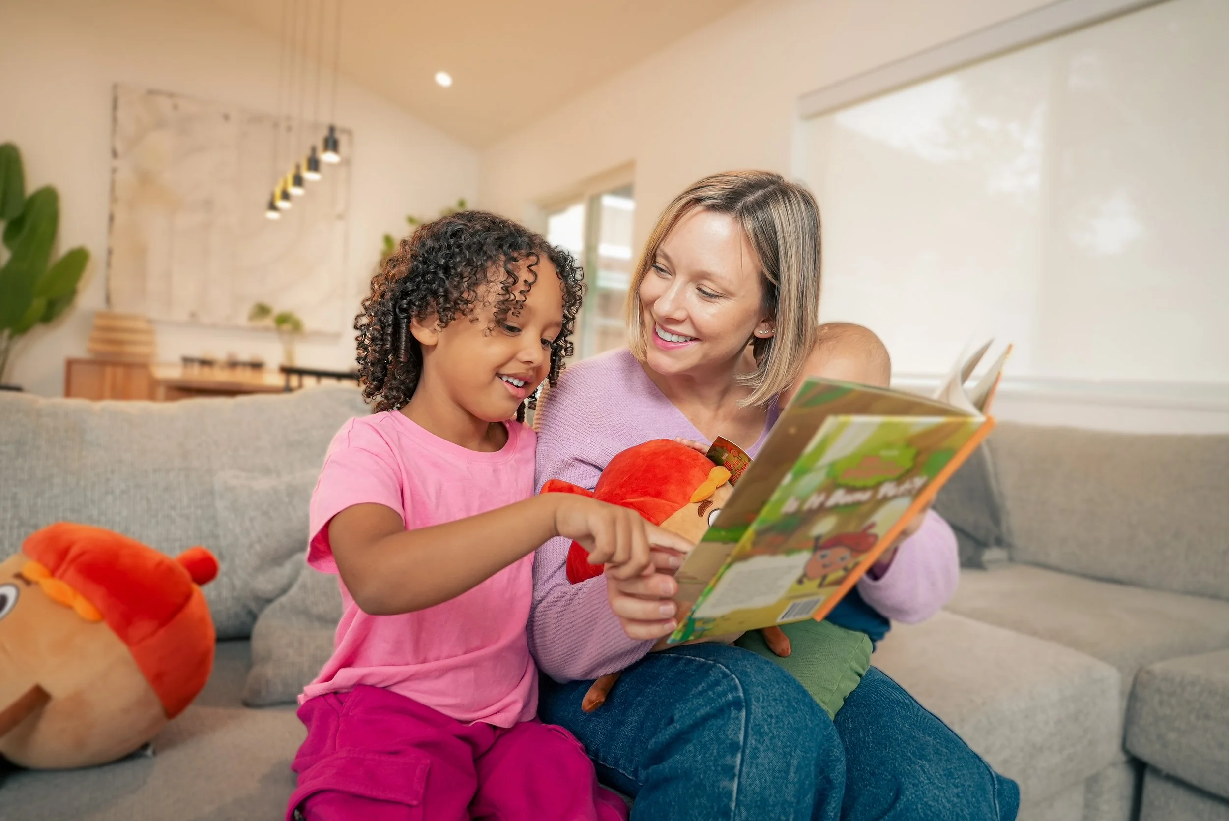 A woman and young girl sitting on a gray couch reading a children's book together, with plush toys around them in a bright living room.