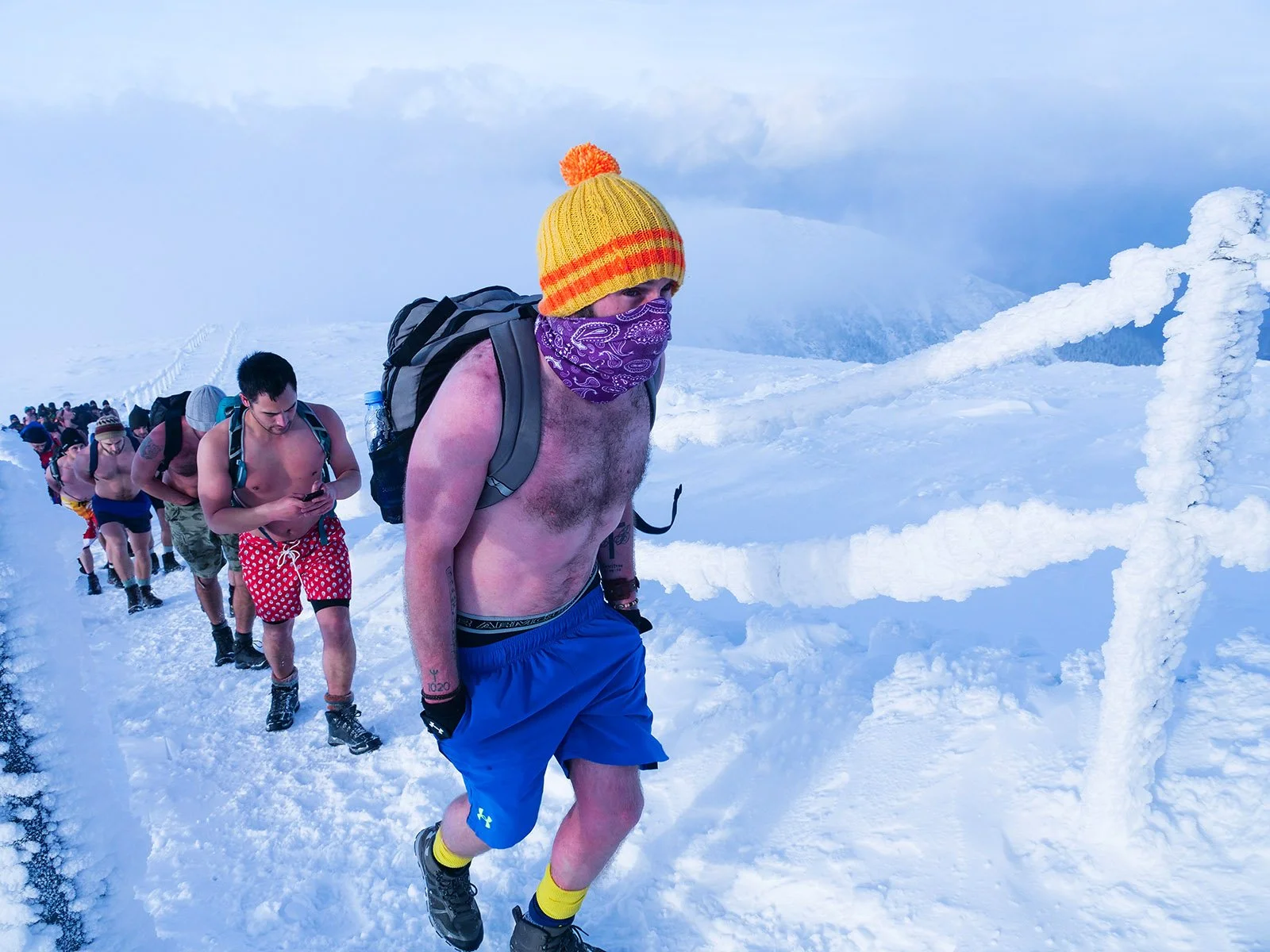 A group of shirtless men with backpacks hiking in a snowy mountain landscape, some wearing colorful winter hats.