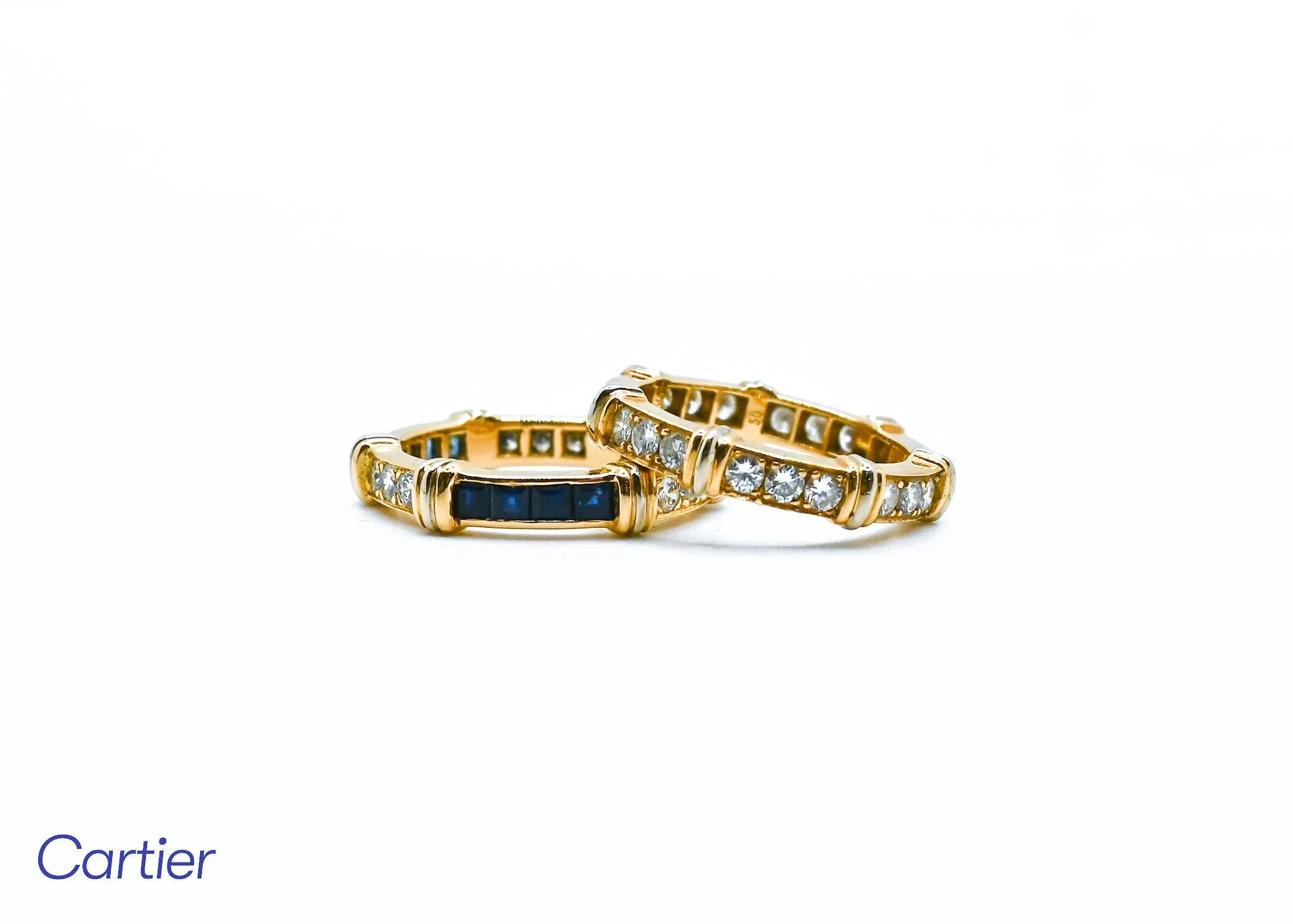 Two gold rings with embedded diamonds, one with black inlays, placed on a white surface with the Cartier logo partially visible in the bottom left corner.