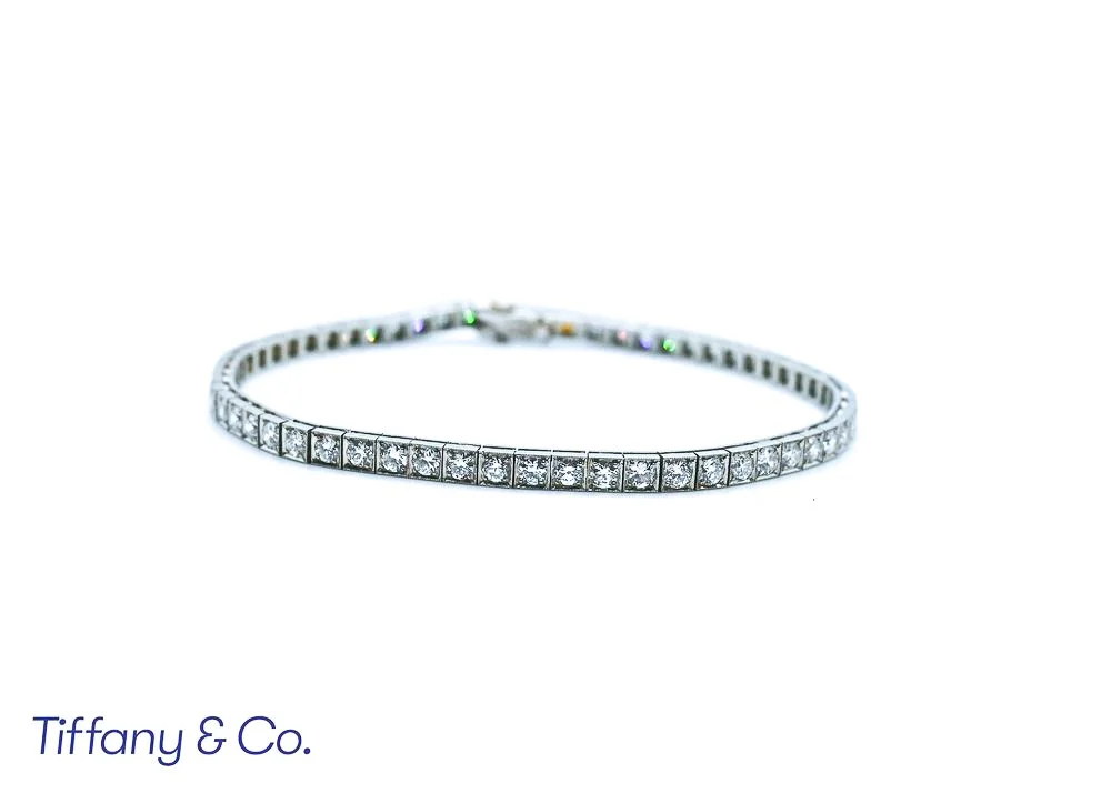 A silver tennis bracelet with square-cut clear stones on a white background, partially visible Tiffany & Co. branding at the bottom left.