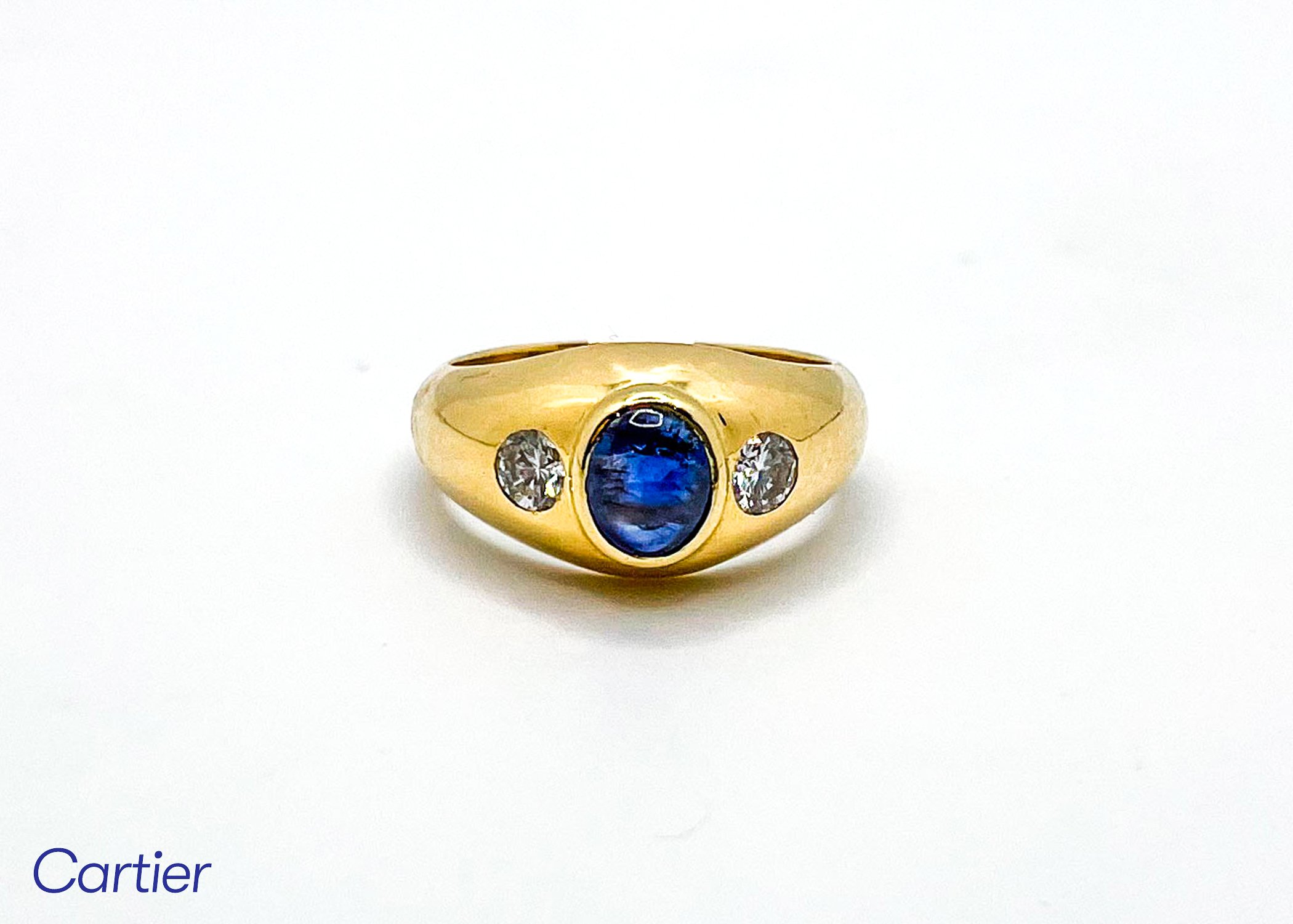 Close-up of a gold ring featuring a large blue gemstone at the center and two smaller clear gemstones on either side, against a white background.
