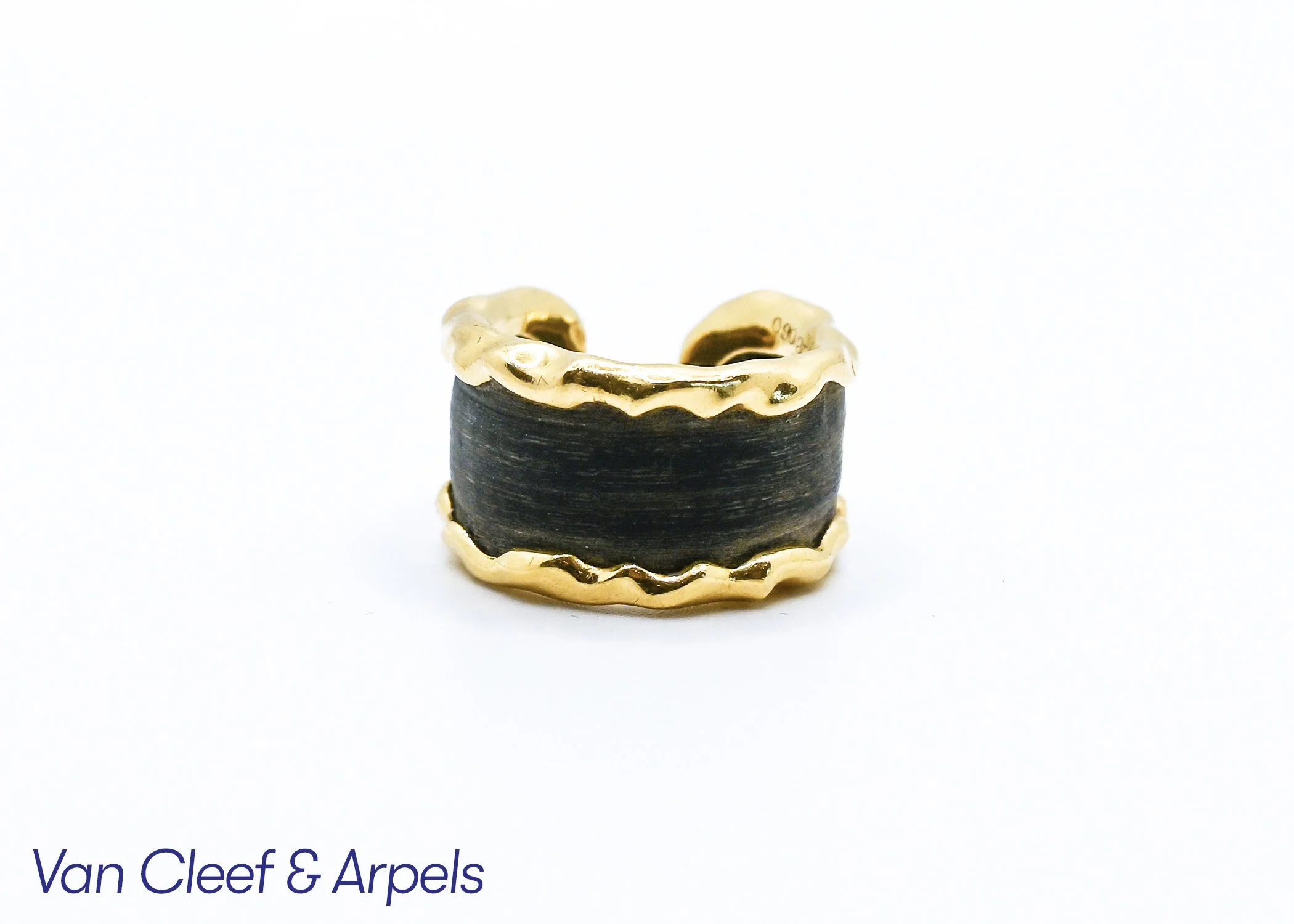 Close-up of a gold and black ring with an elegant, wavy design against a white background, with partial text reading 'Van Cleef & Arpels' at the bottom.