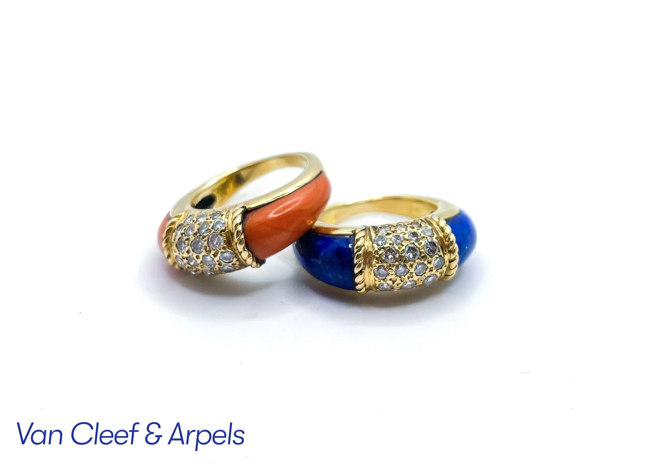 Two gold rings, each decorated with diamonds and colorful enamel. One ring with orange enamel and the other with blue enamel, resting on a white background with partial text reading 'Van Cleef & Arpels'.