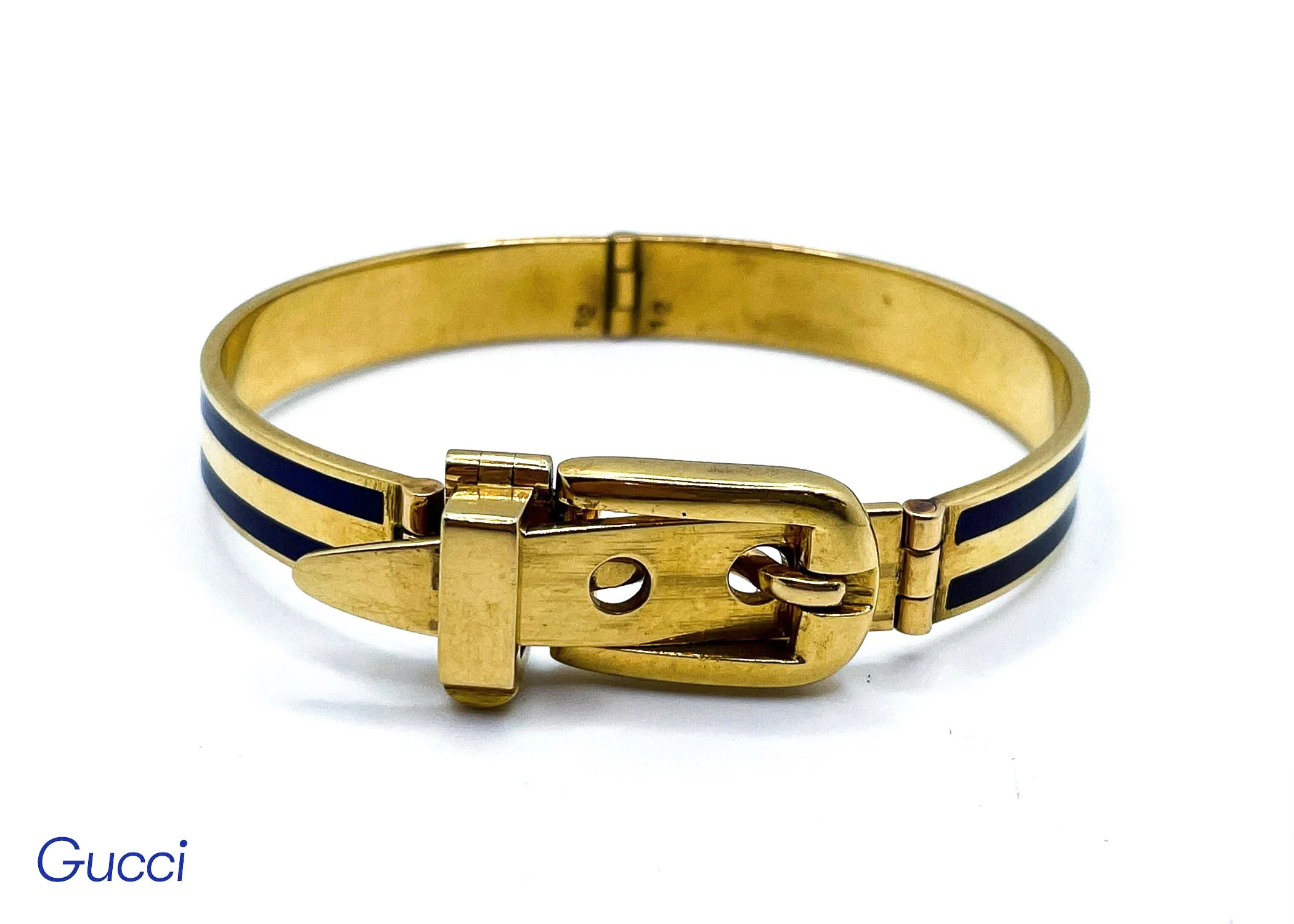 A gold Gucci bracelet with black accents and a gold clasp placed on a white surface with the Gucci logo partially visible.