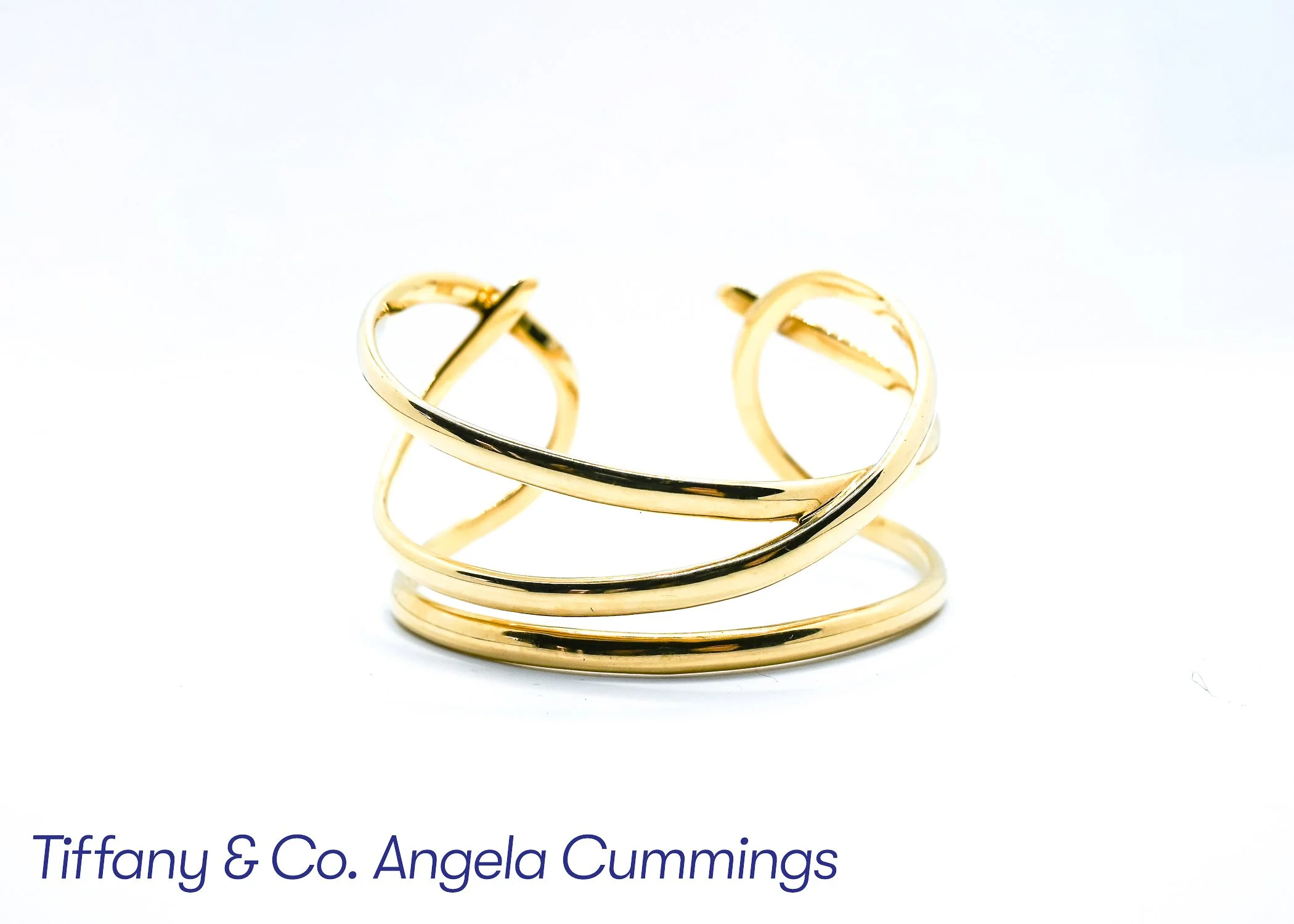 Close-up of multiple gold rings stacked together in the foreground with the text "Tiffany & Co. Angela Cummings" at the bottom.
