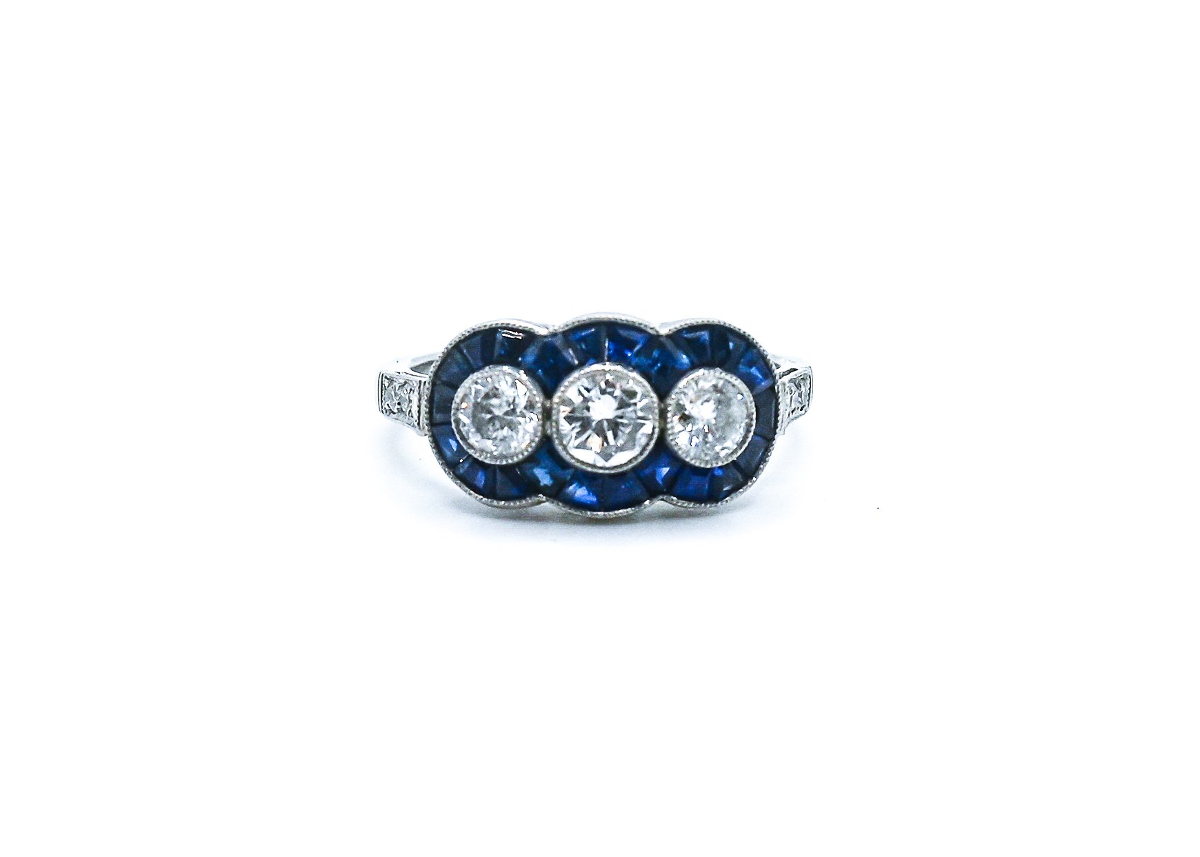 A silver ring with three large central diamonds surrounded by blue gemstones and smaller diamonds on the band, set against a white background.