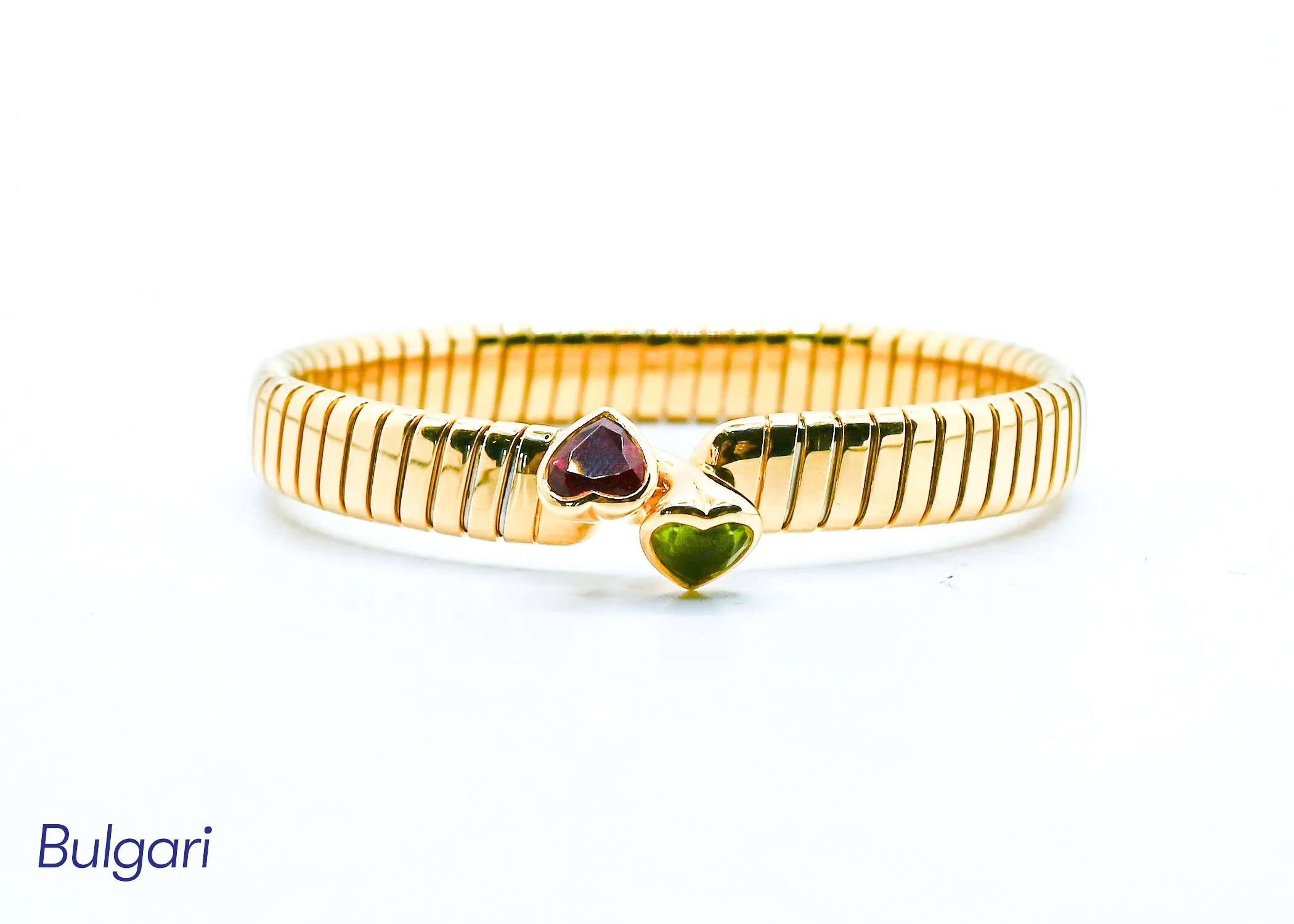 A gold jewelry ring with a heart-shaped purple gemstone and a green gemstone, displayed against a white background with partially visible text 'Bulgari' in the lower left corner.