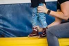 A child in blue jeans with cartoon characters is standing on a yellow padded mat. An adult, wearing a black shirt and blue jeans, is kneeling and holding the child's foot, helping them put on a purple and pink sneaker.