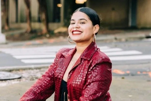 A smiling woman with dark hair, wearing hoop earrings and a red textured jacket, sitting outdoors near a street with trees in the background.