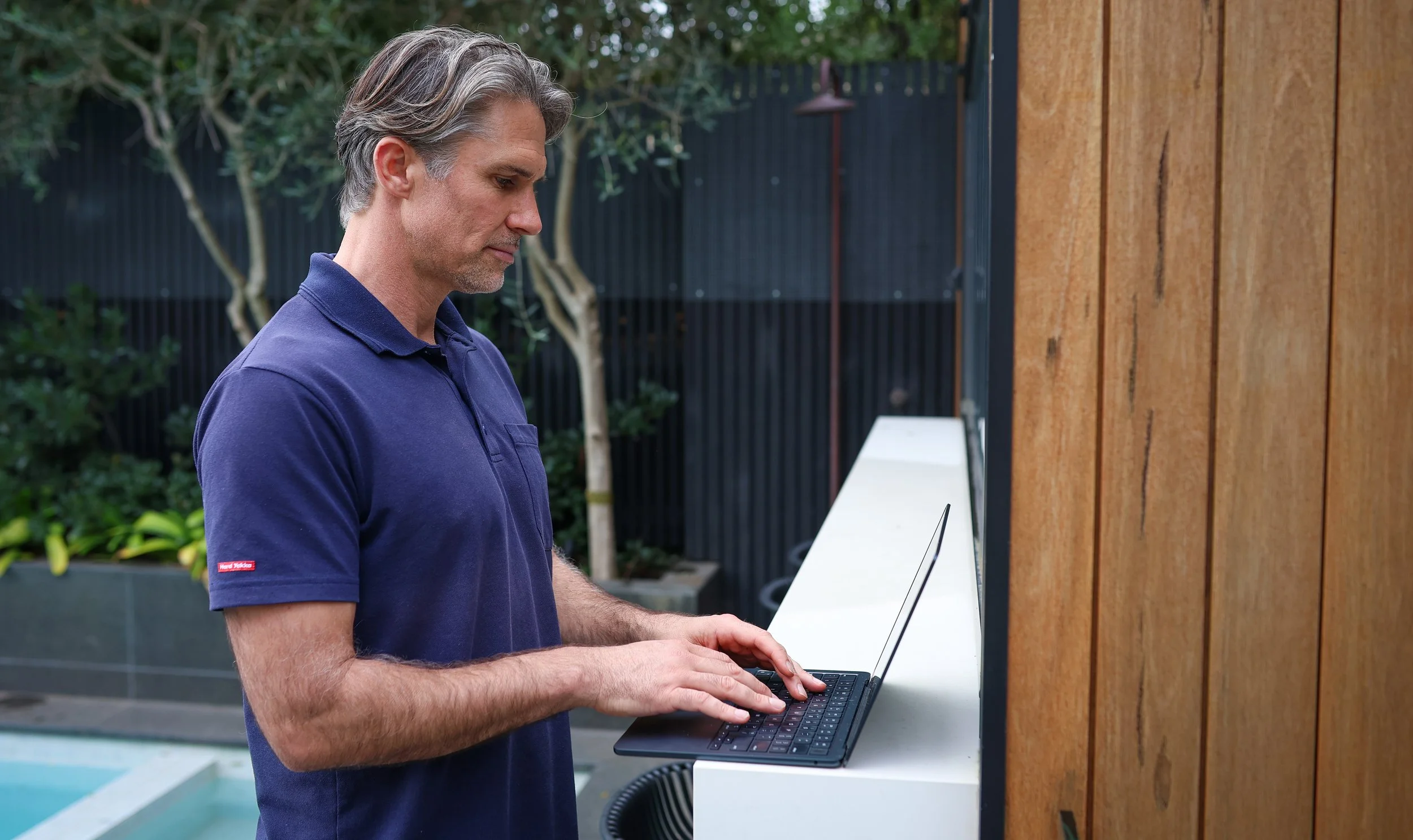 A middle-aged man in a blue polo shirt using a laptop outdoors beside a wooden wall.