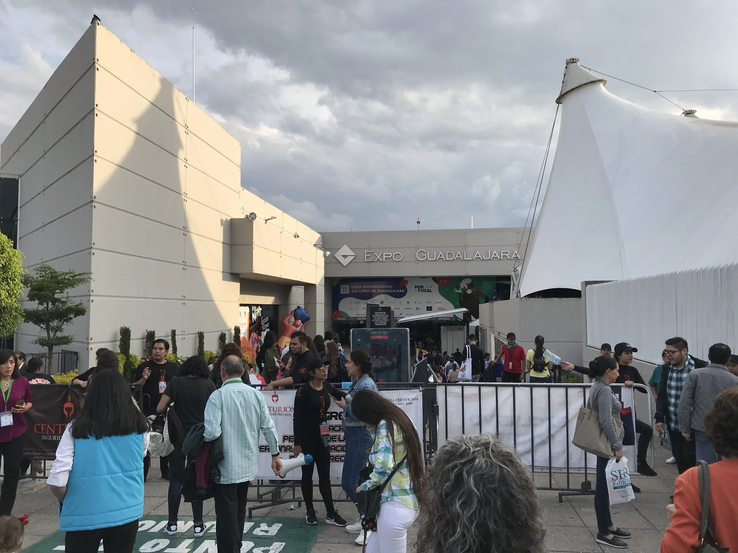 Crowd of people in front of Expo Guadalajara entrance, some talking, some holding bags, under a cloudy sky, with the expo building visible behind.