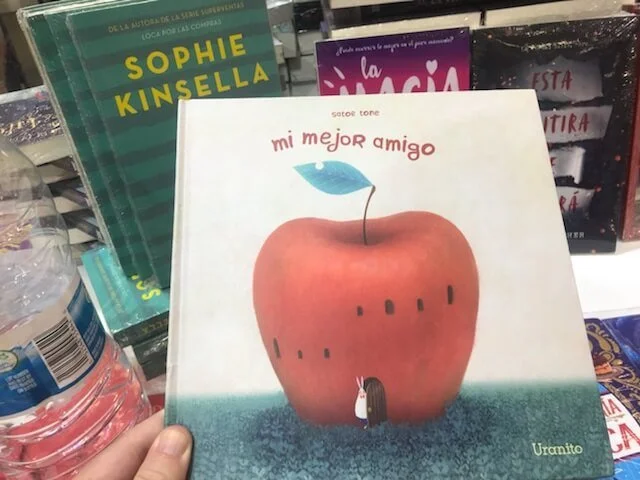 A book titled 'Mi mejor amigo' with an illustration of an apple with a door and a small character standing in front. The background includes other books and a water bottle.