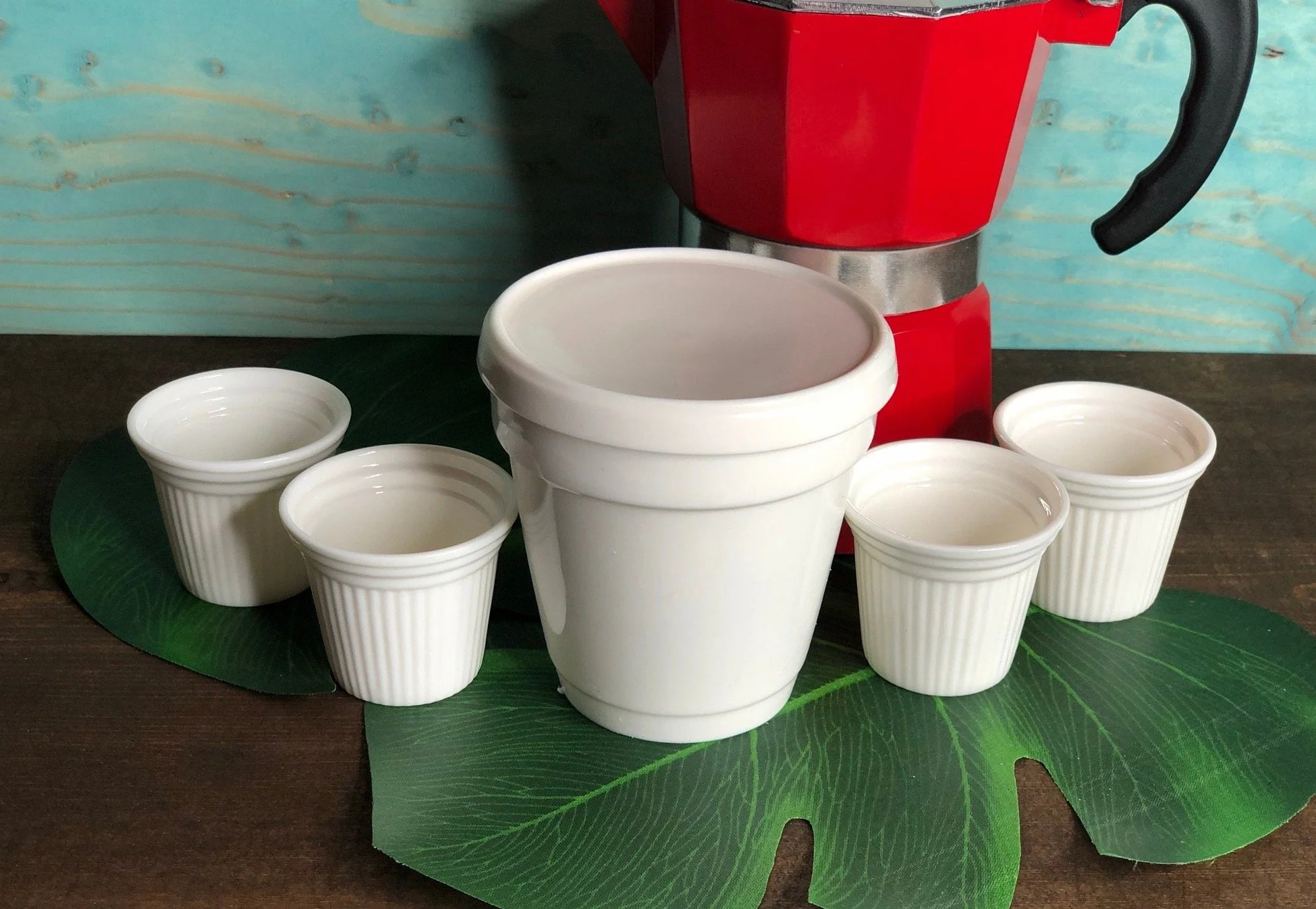 Four small white ceramic espresso cups, one large white ceramic cup, and a decorative moka coffee maker on a green tropical leaf placemat, with a blue wooden background.