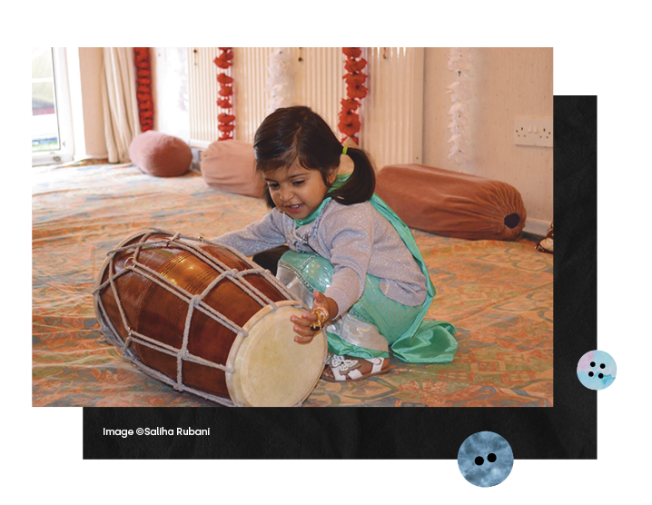  A very young girl in a colourful kneels down to play with large drum instrument and a young woman makes notes intently in front of images hung in a gallery 